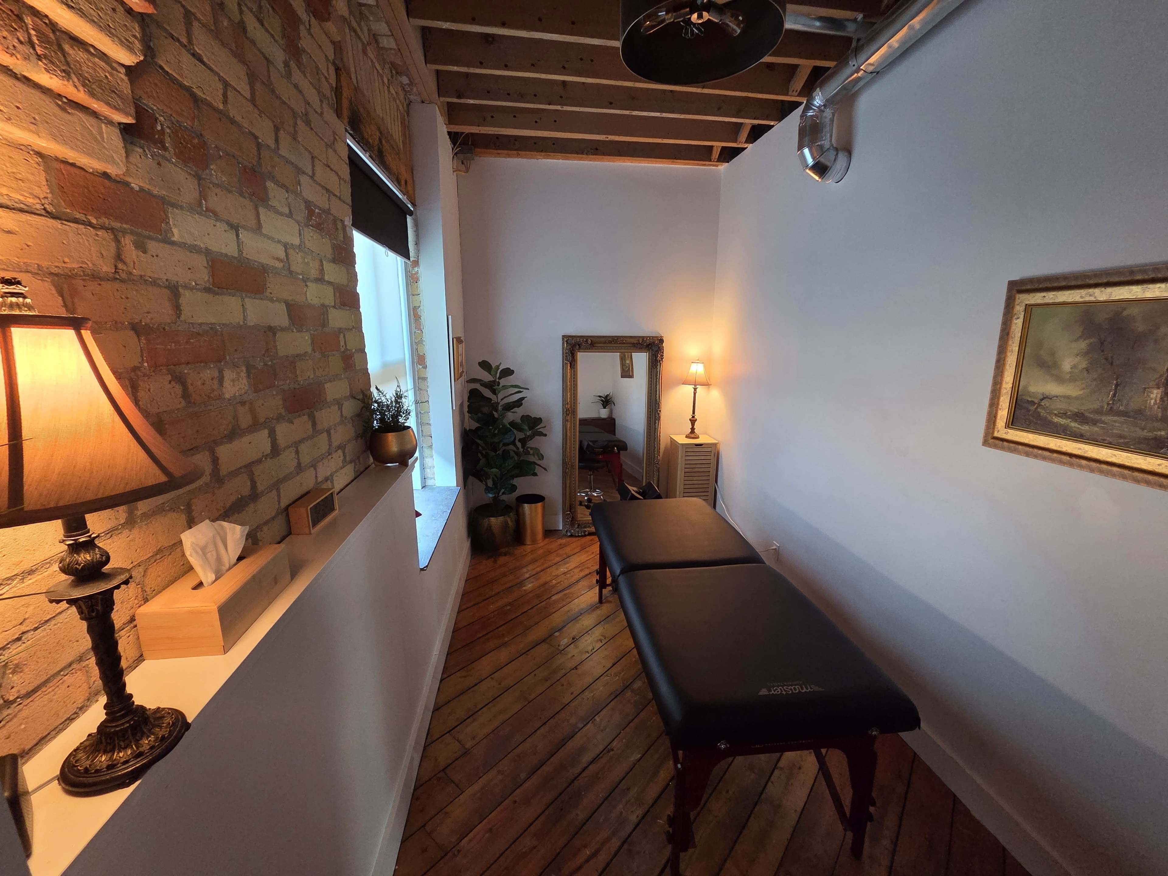 The image shows a small massage therapy room with brick walls, wooden floors, and a massage table centered in the space, accompanied by a lamp and a mirror.