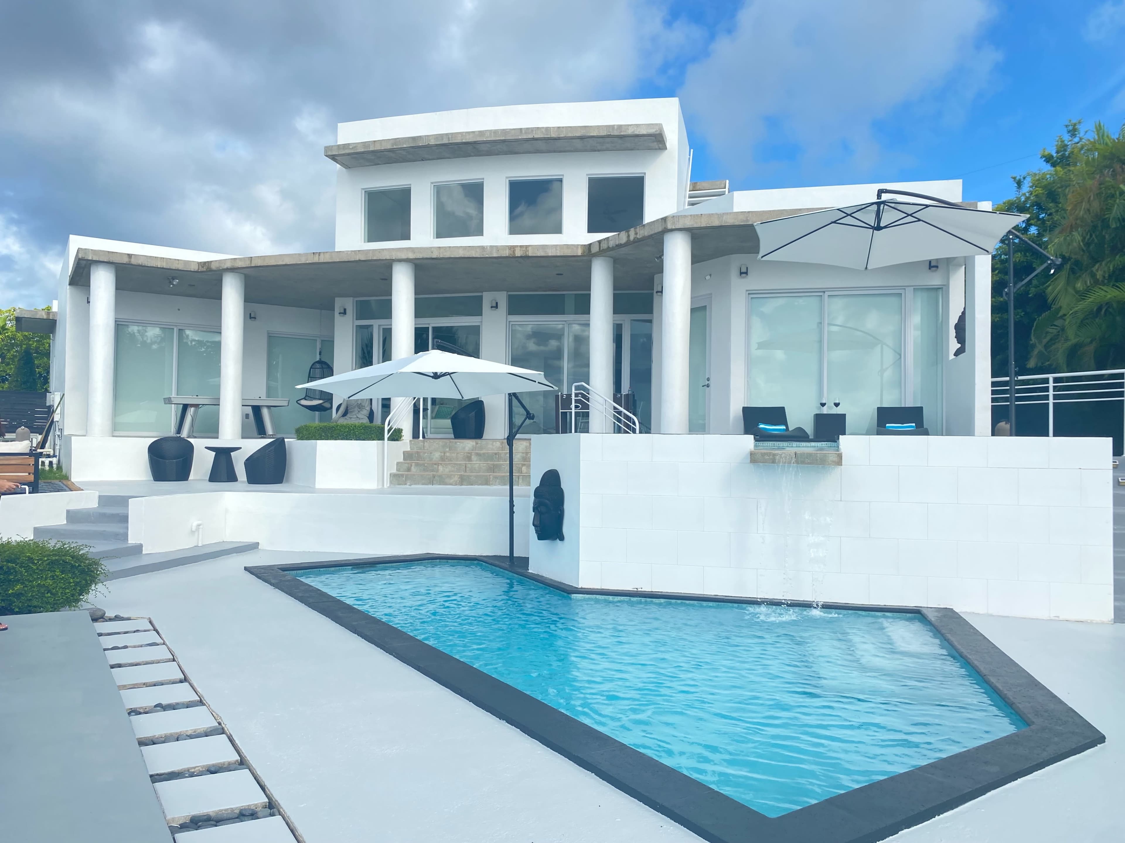 A modern white house with large windows, a small pool in front, and an umbrella over outdoor seating areas.