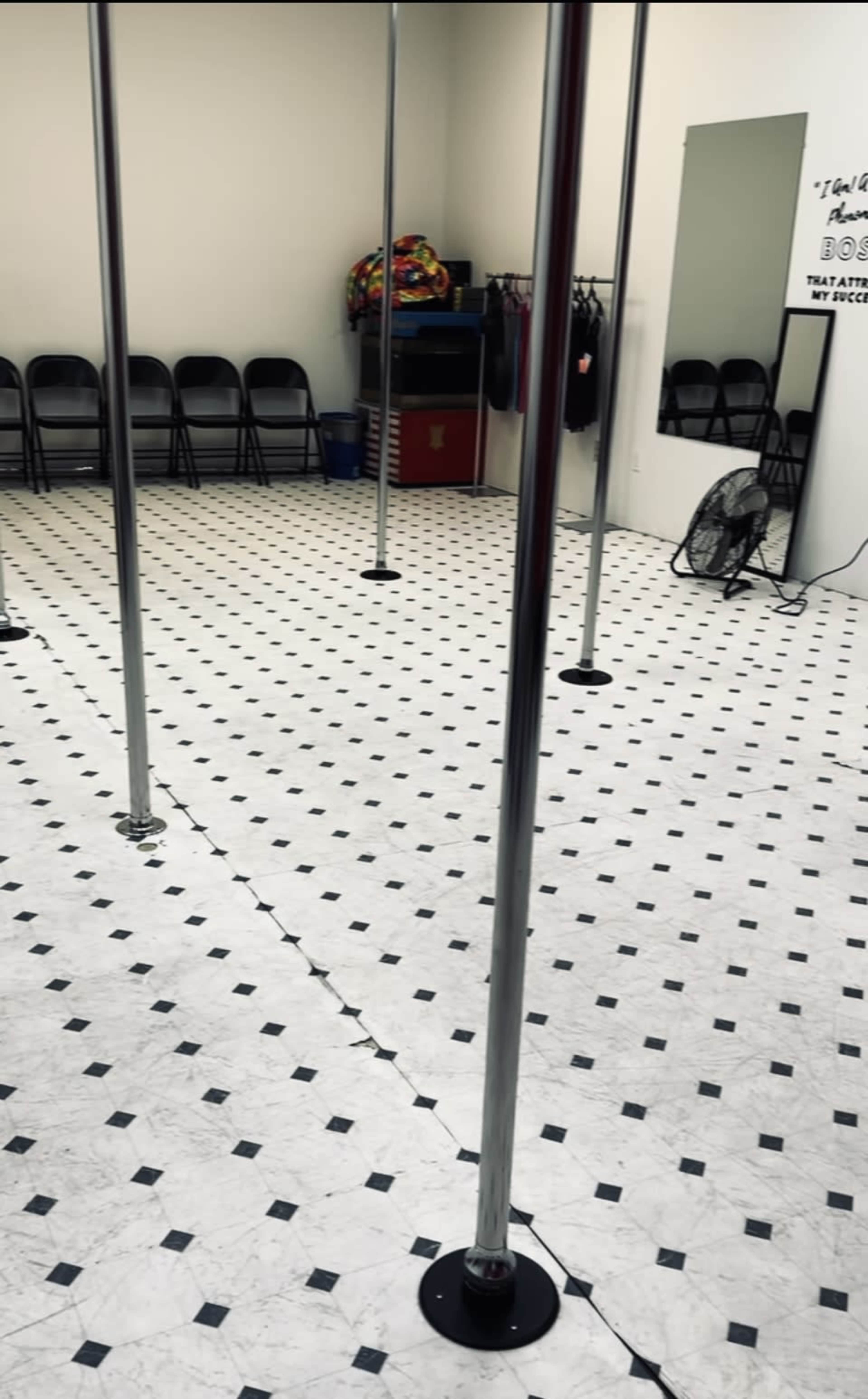The image shows an empty room with multiple shiny pole dancing poles on a tiled floor, surrounded by folding chairs and a mirror along one wall.