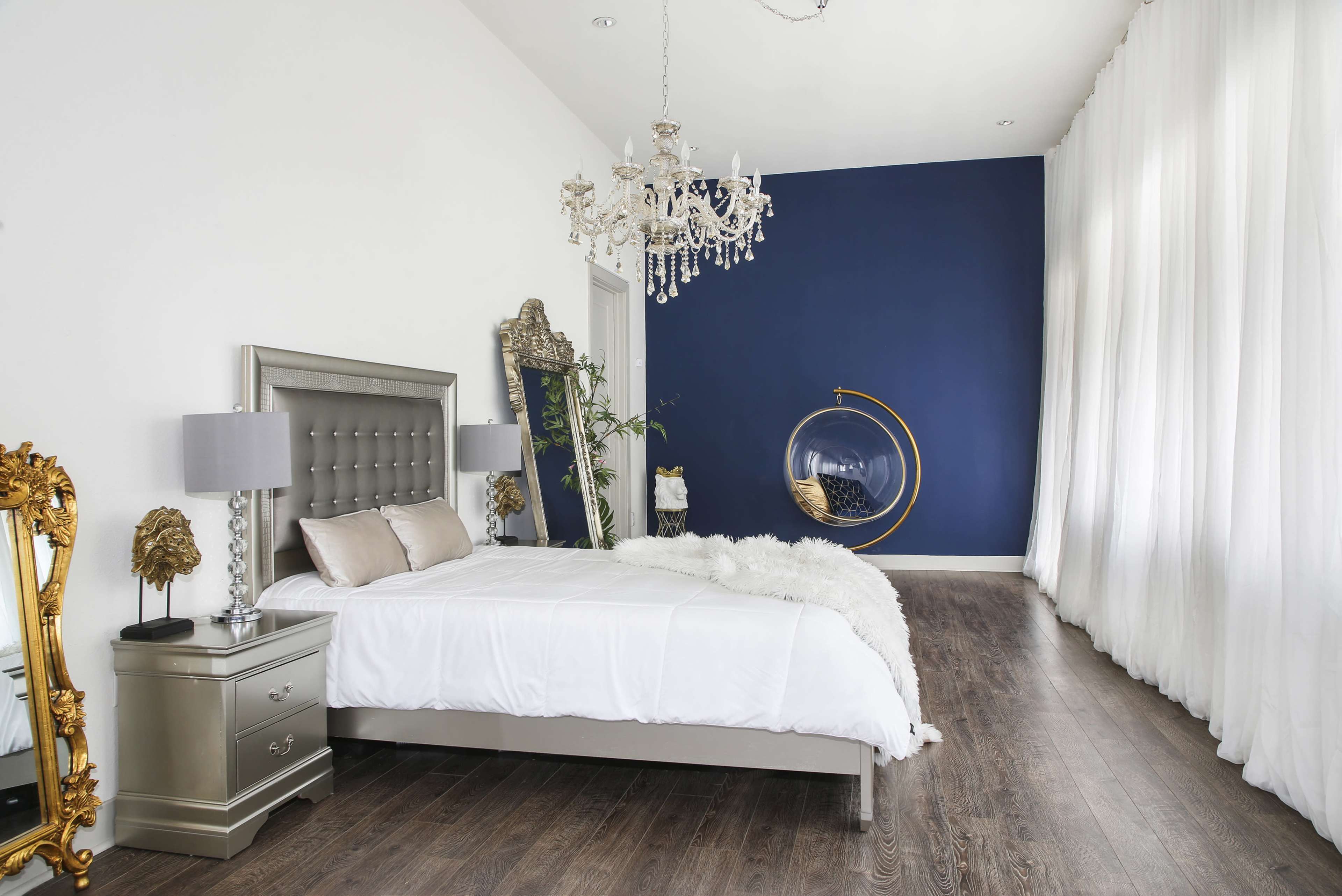 The image shows a modern bedroom featuring a silver bed, large windows with white curtains, a blue accent wall, and a decorative chandelier.
