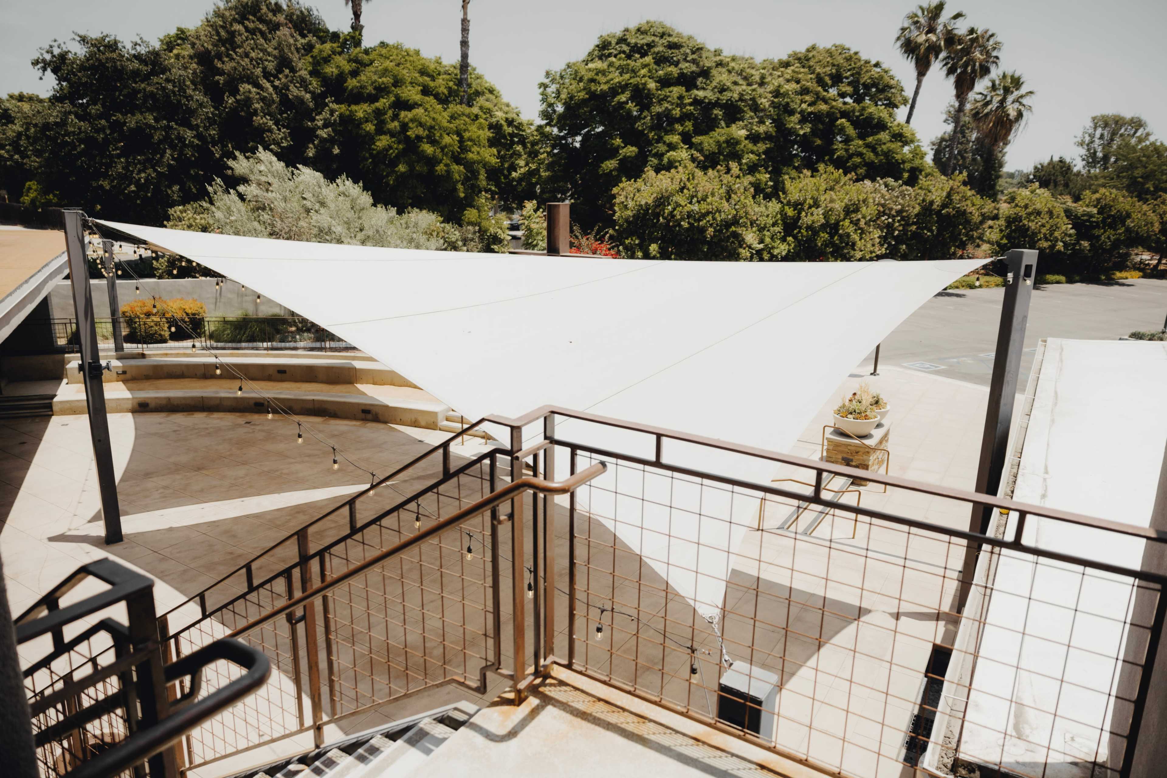 A white shade sail is installed over a patio area, supported by metal poles, with trees and foliage in the background.
