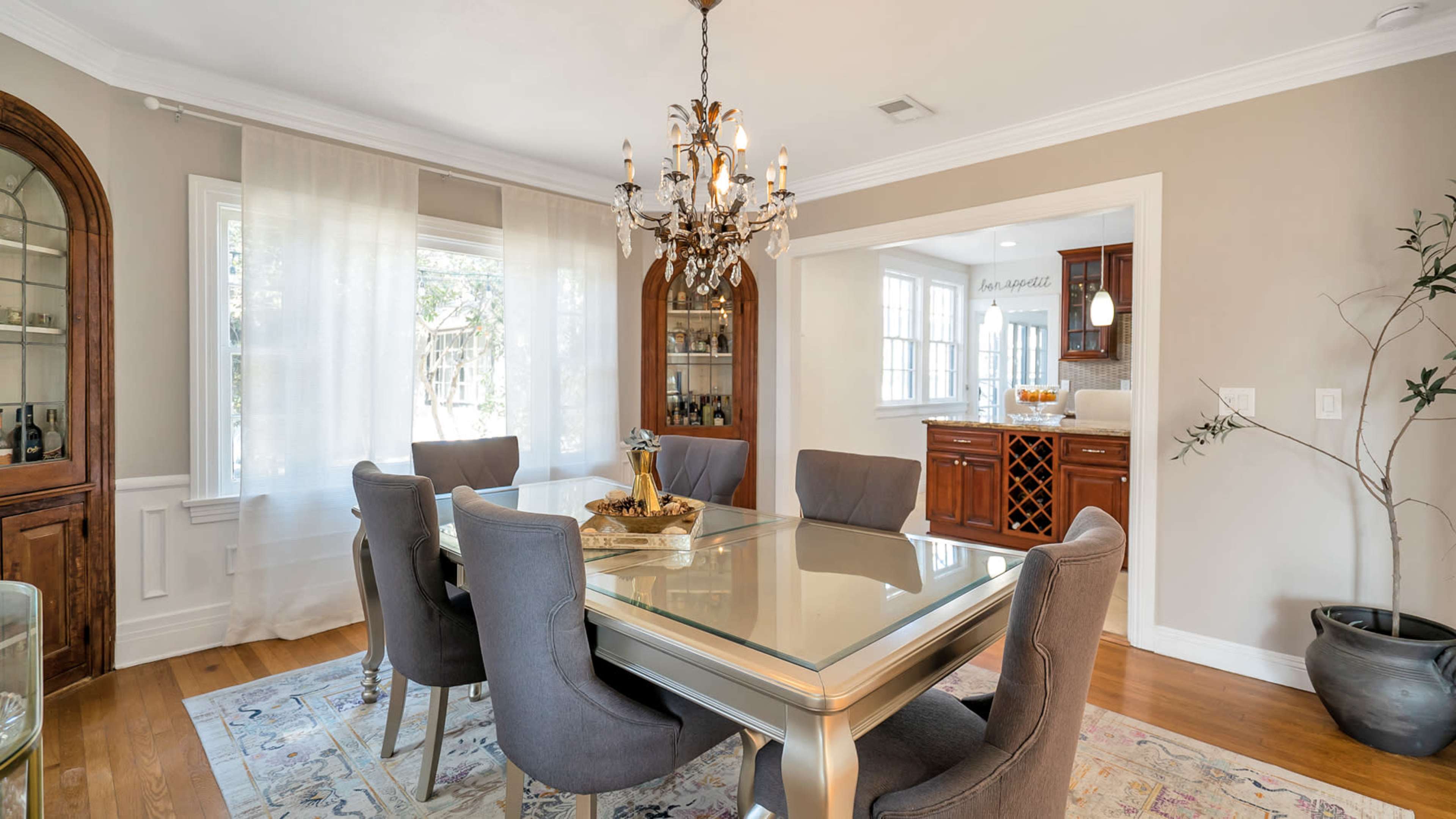 A dining room features a glass-topped table surrounded by upholstered chairs, with a chandelier overhead and a doorway leading to a kitchen.