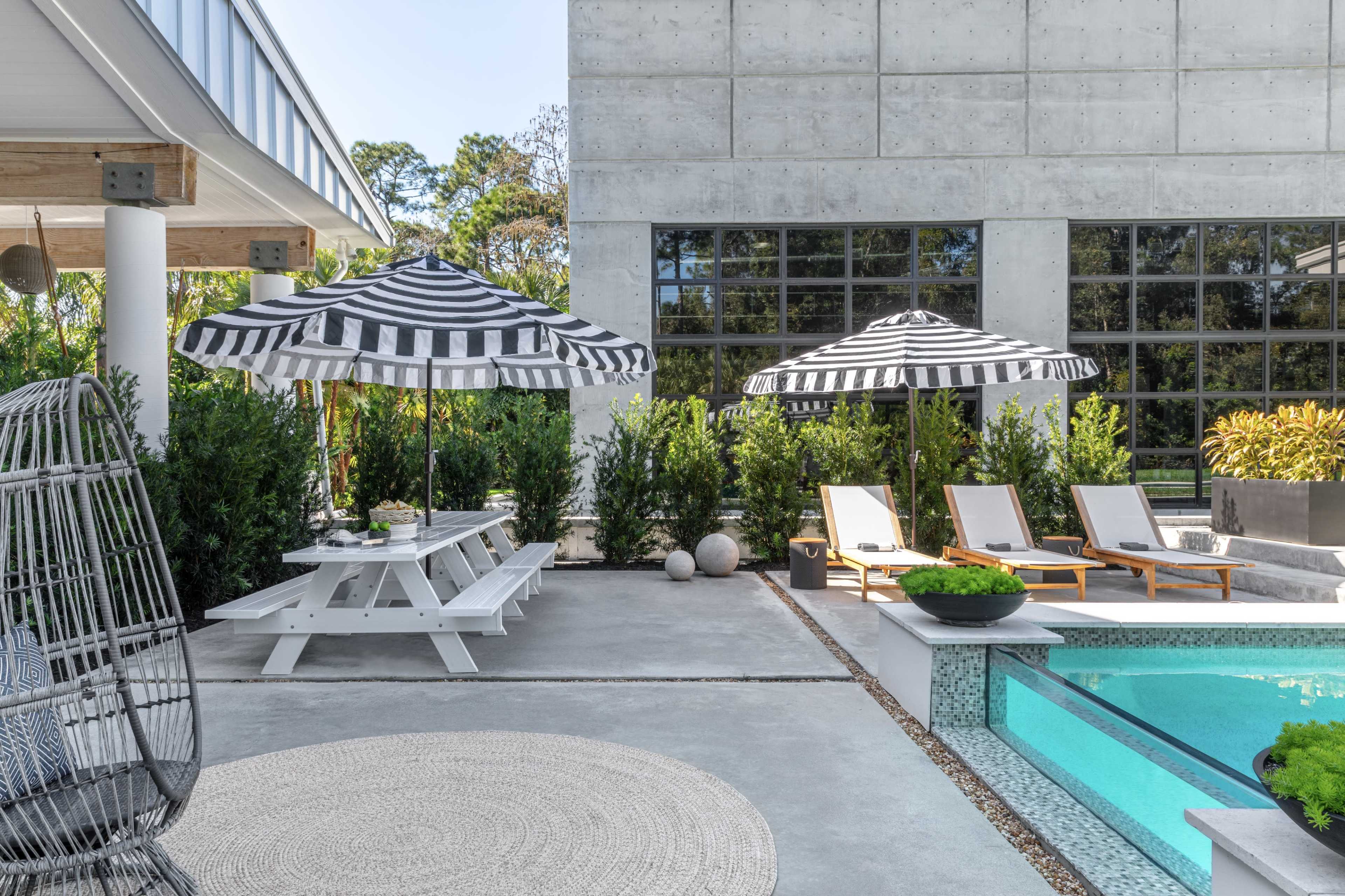 The image features a modern outdoor space with a concrete patio, a swimming pool, two striped umbrellas over lounge chairs, and a picnic table.