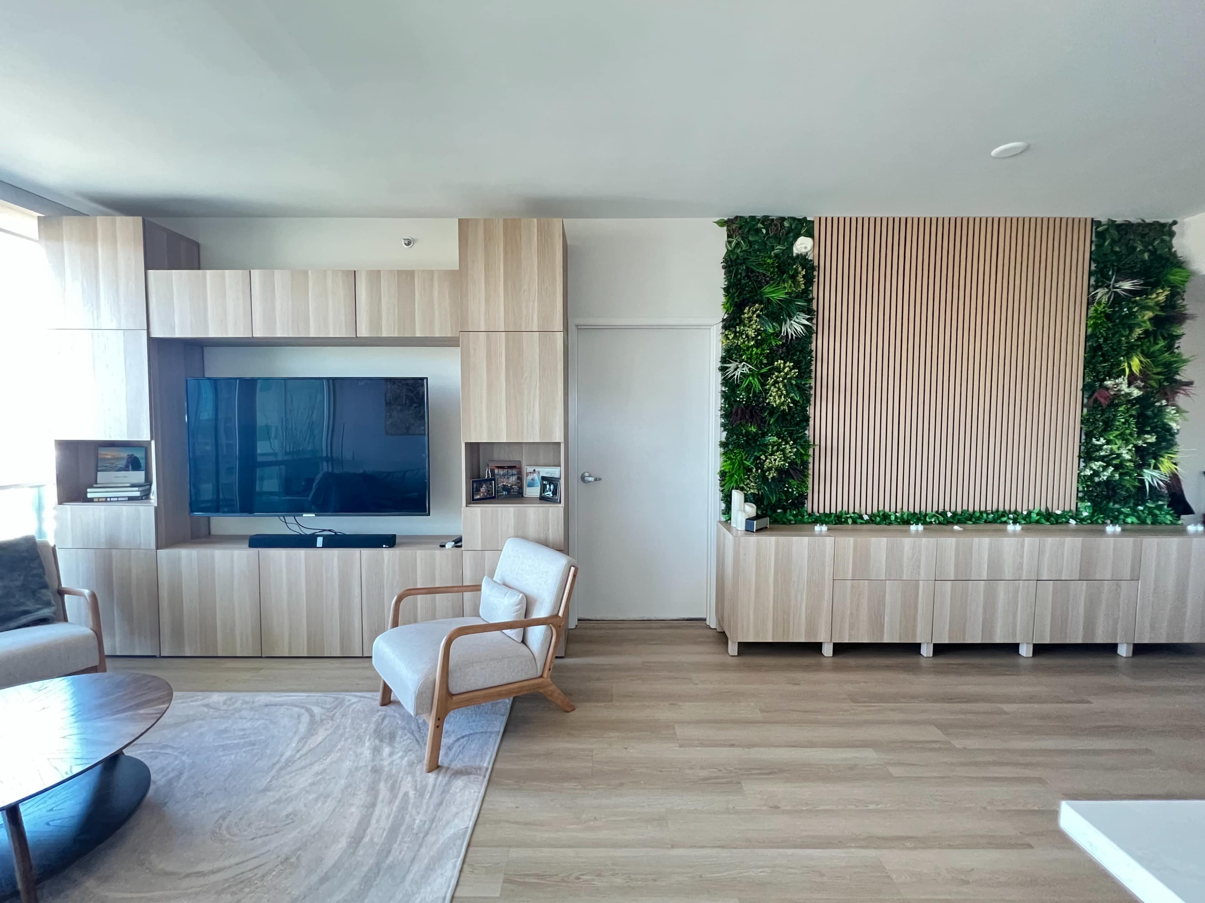 The image shows a modern living room with light wood furniture, a TV mounted on a feature wall, and a vertical garden on another wall.