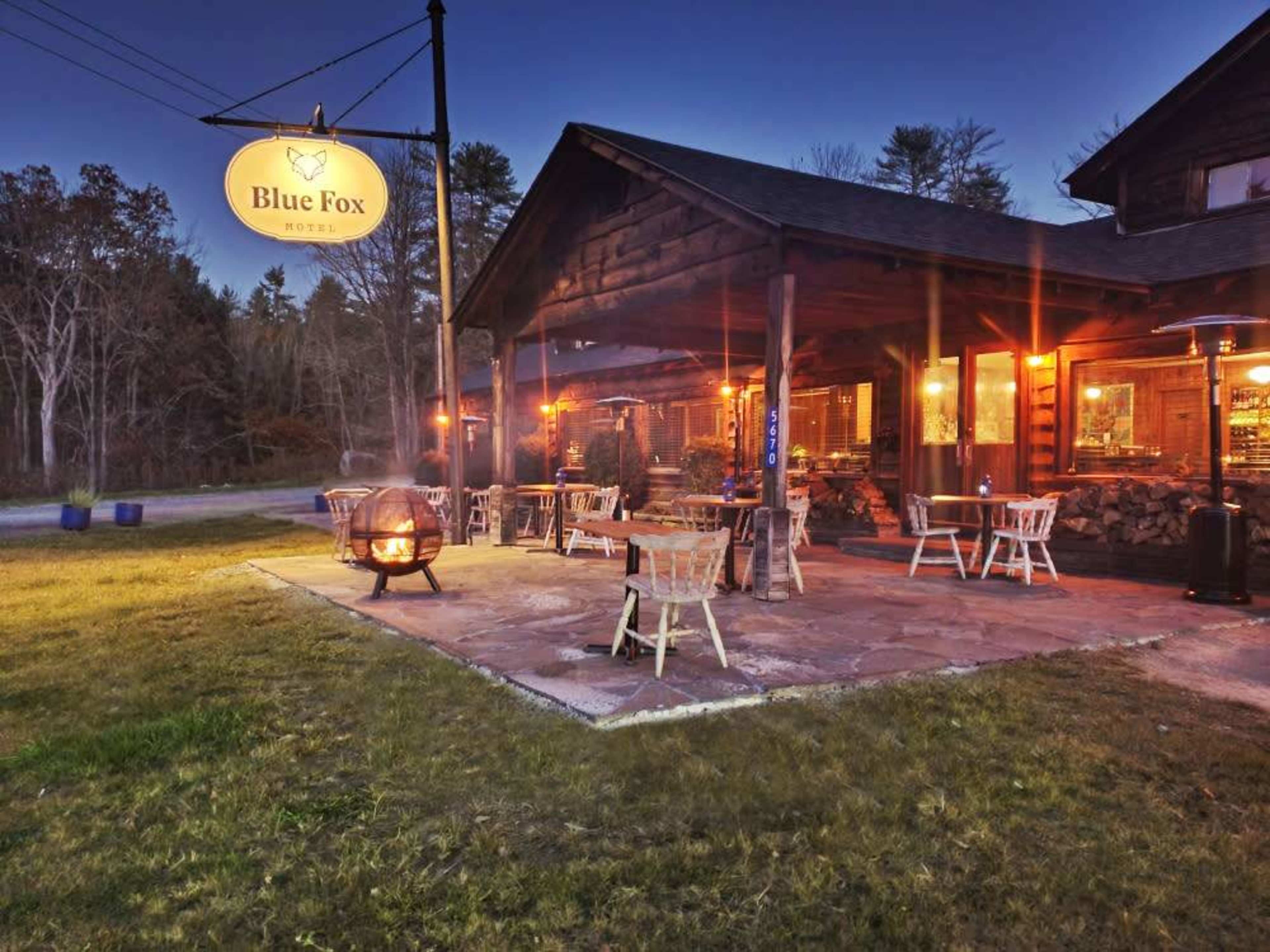 A rustic lodge called "Blue Fox Motel" with outdoor seating illuminated by warm lights on a grassy area at dusk.