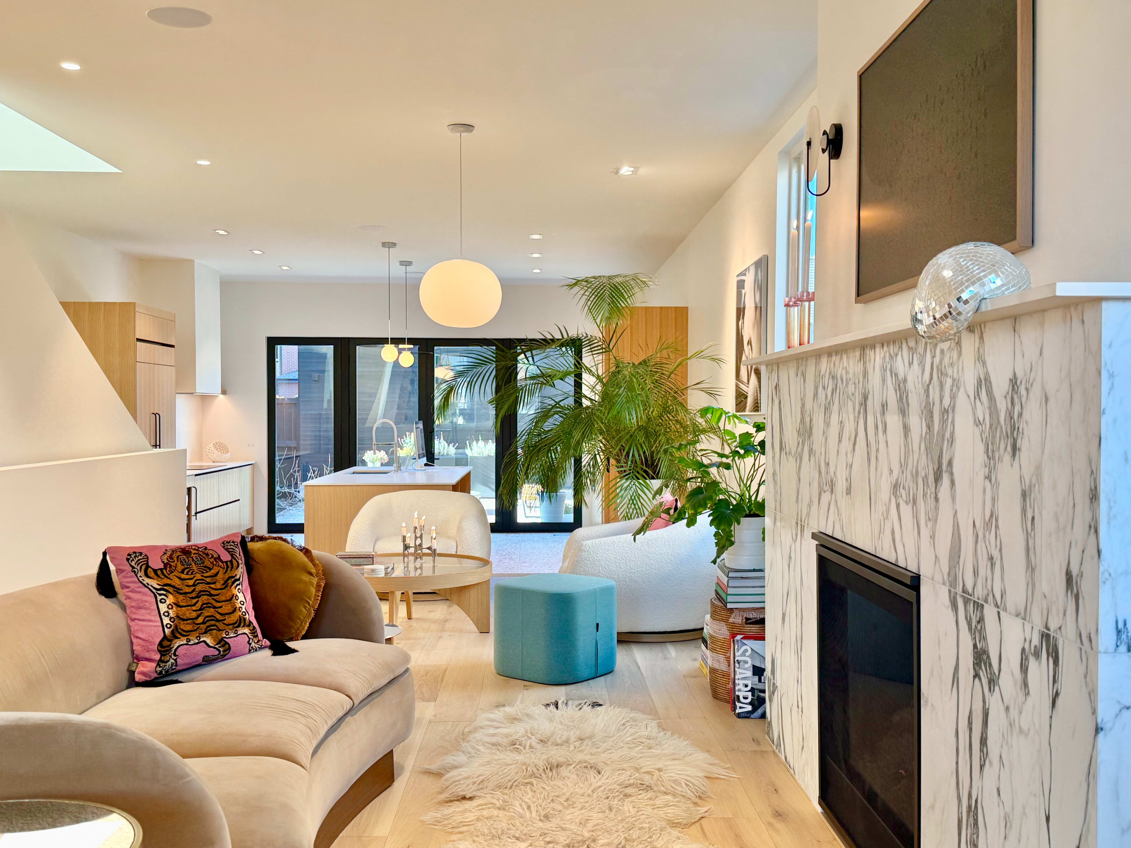 The image shows a modern living room with a light wooden floor, a marble fireplace, a large window, and contemporary furniture including a pink sofa and a blue ottoman.