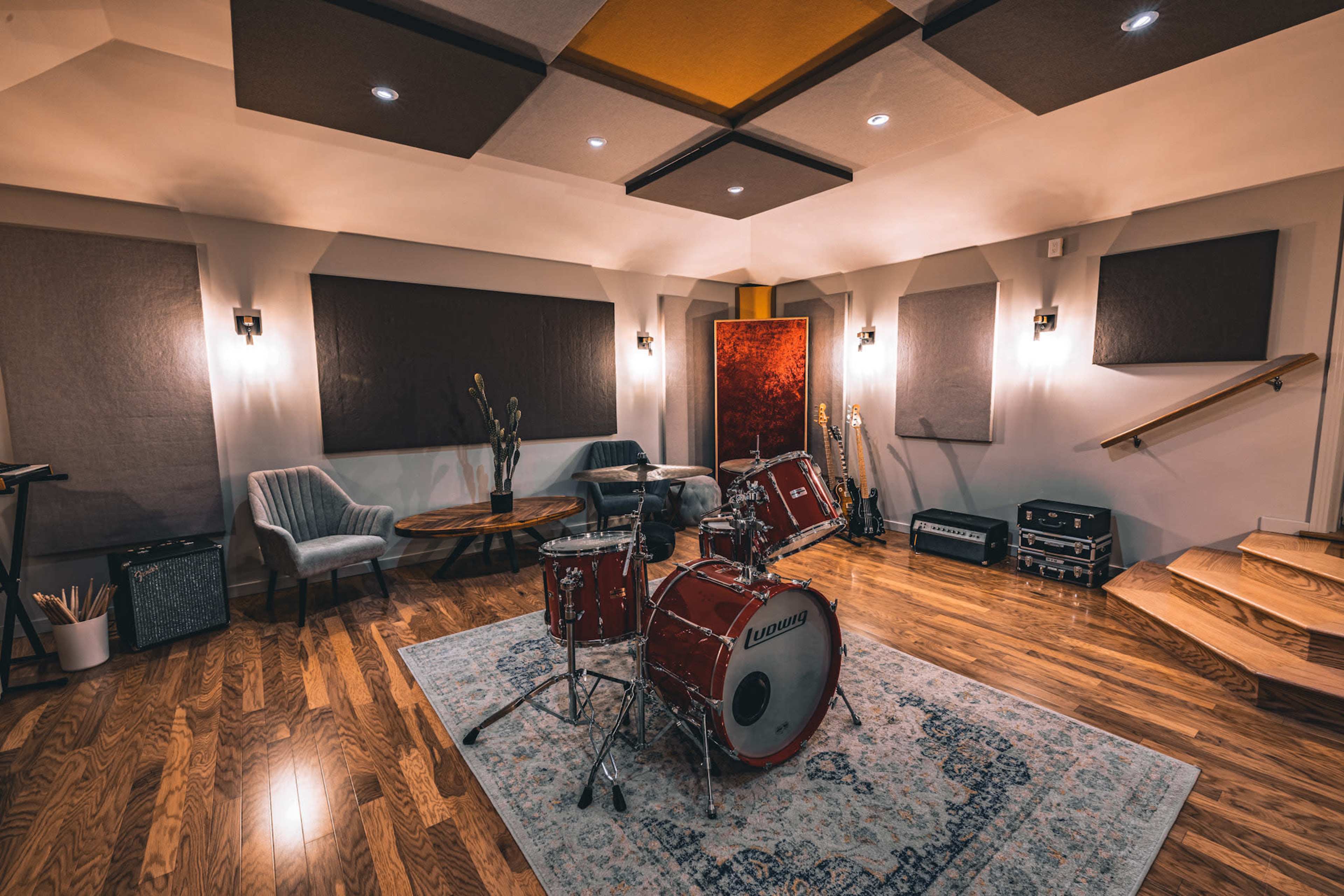 A music studio featuring a red drum set on a patterned rug, surrounded by wooden flooring, acoustic panels, and various musical instruments and equipment.