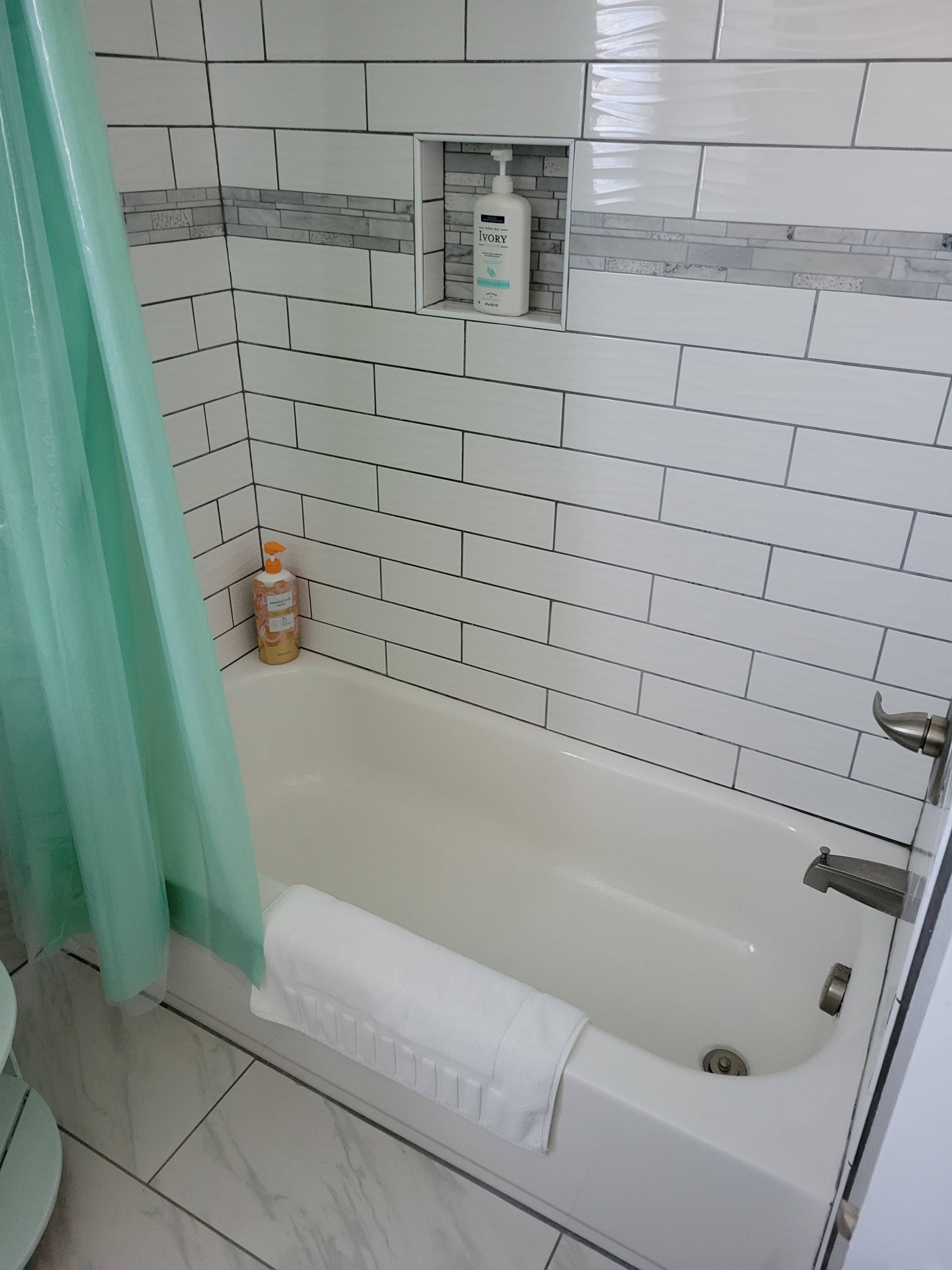The image shows a clean bathroom with a white bathtub surrounded by tiled walls, a light green shower curtain, and a small shelf containing personal care products.