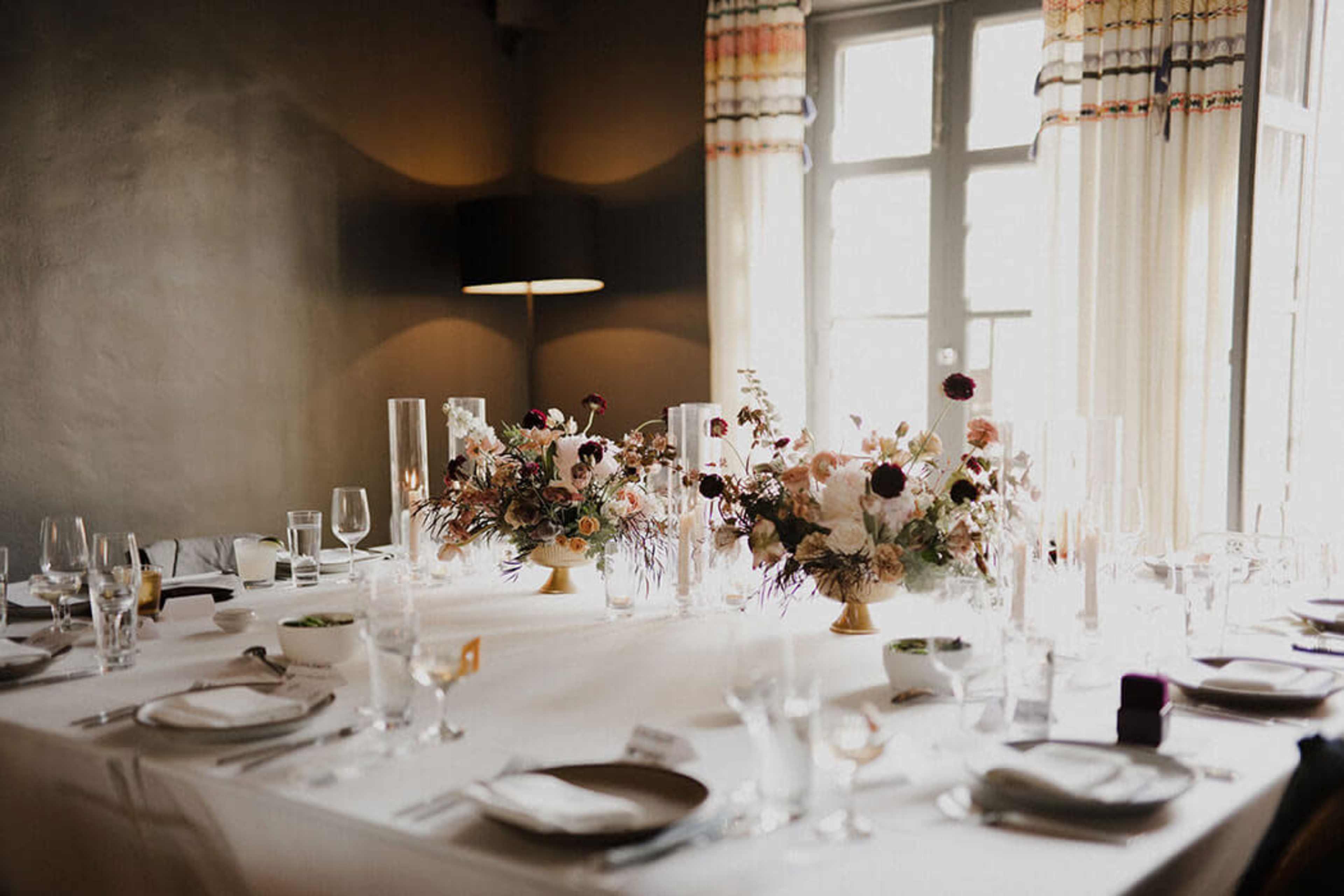 A dining table is elegantly set with plates and glassware, featuring floral centerpieces and soft lighting from a nearby lamp.