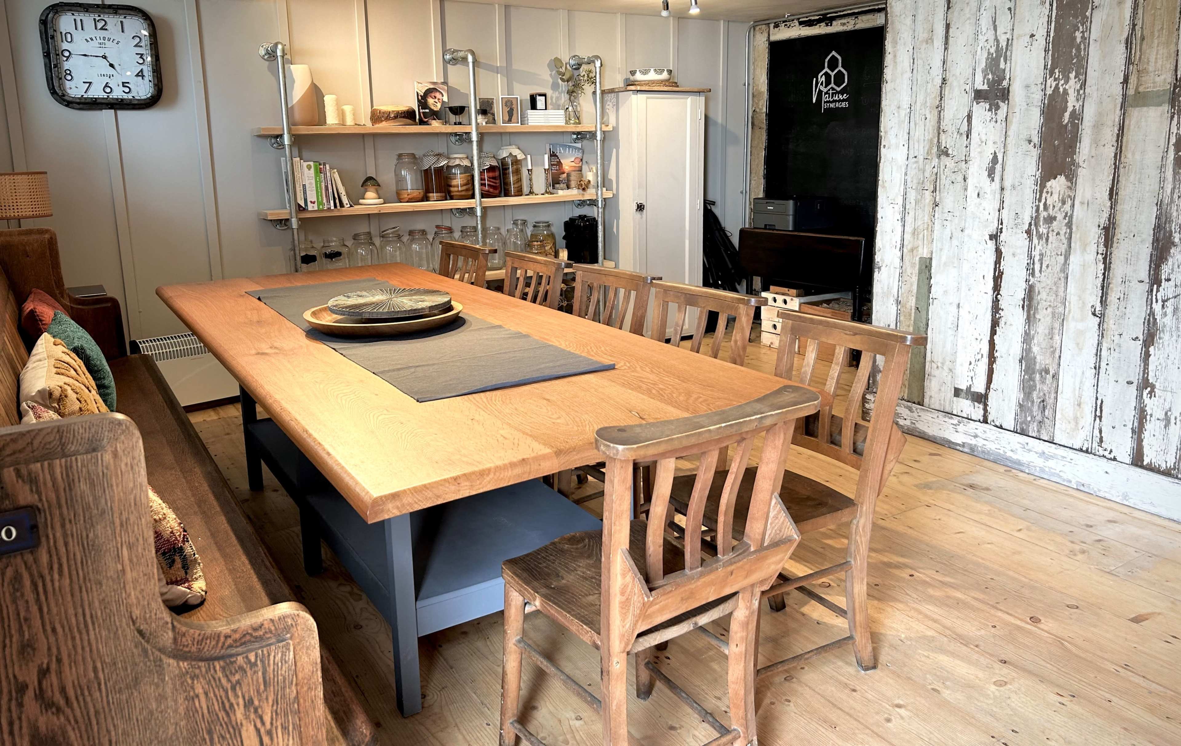 A long wooden dining table surrounded by chairs is set in a rustic room with shelves, a clock, and a chalkboard wall.
