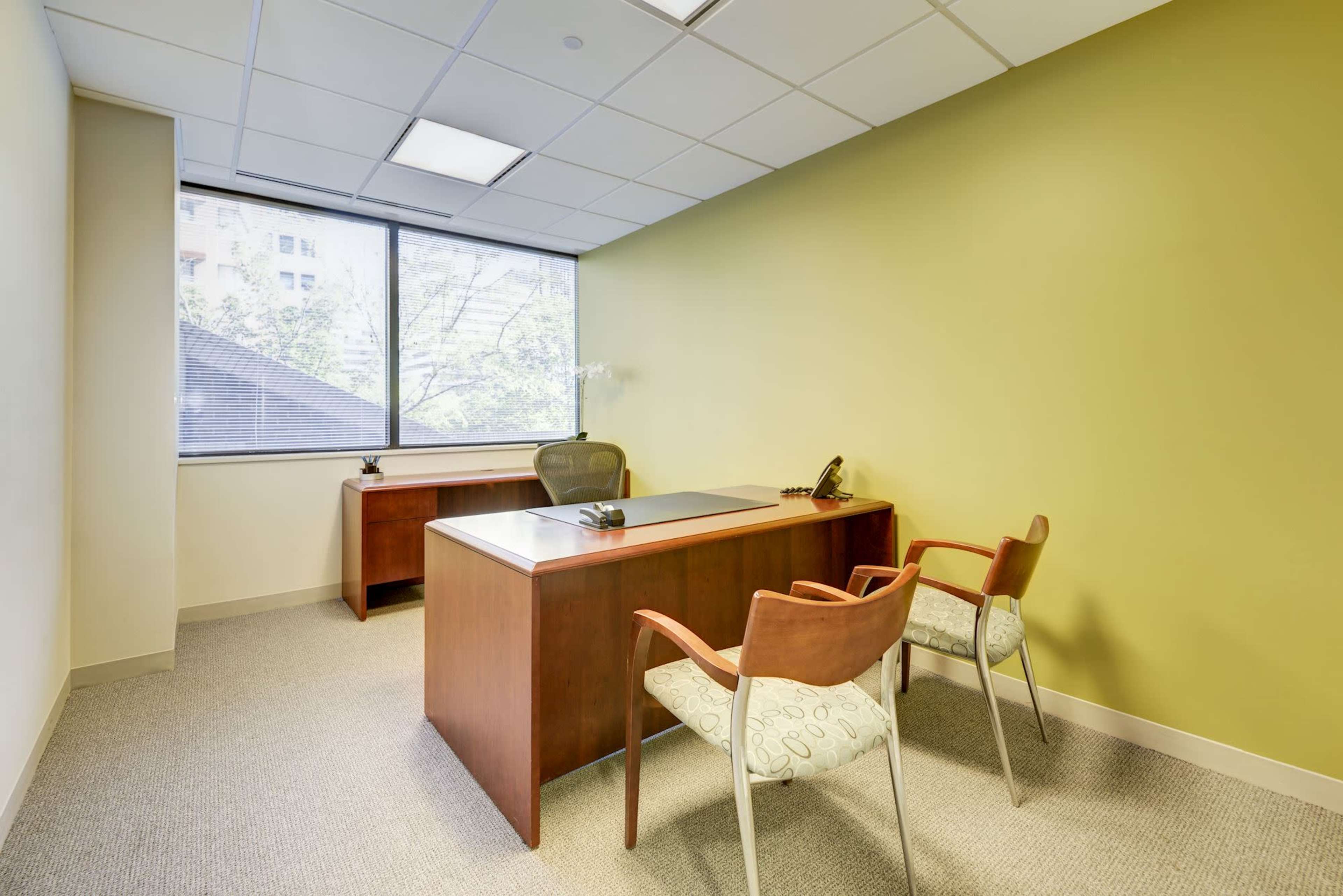 A small office with a wooden desk, an ergonomic chair, and two wooden chairs against a green wall, with a large window providing natural light.