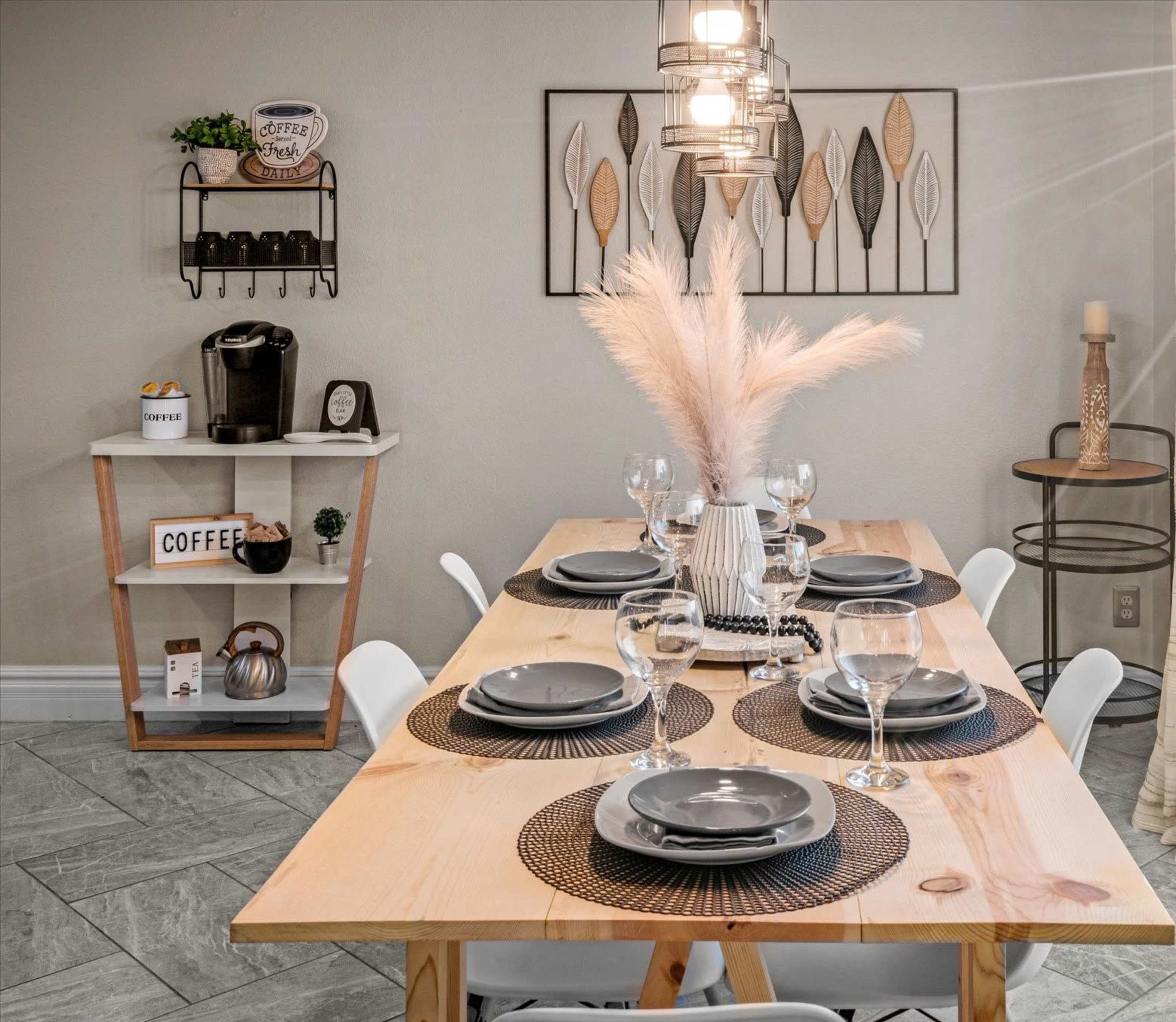 A wooden dining table set with black plates and glasses is arranged in a modern dining area featuring decorative wall art and a coffee station.