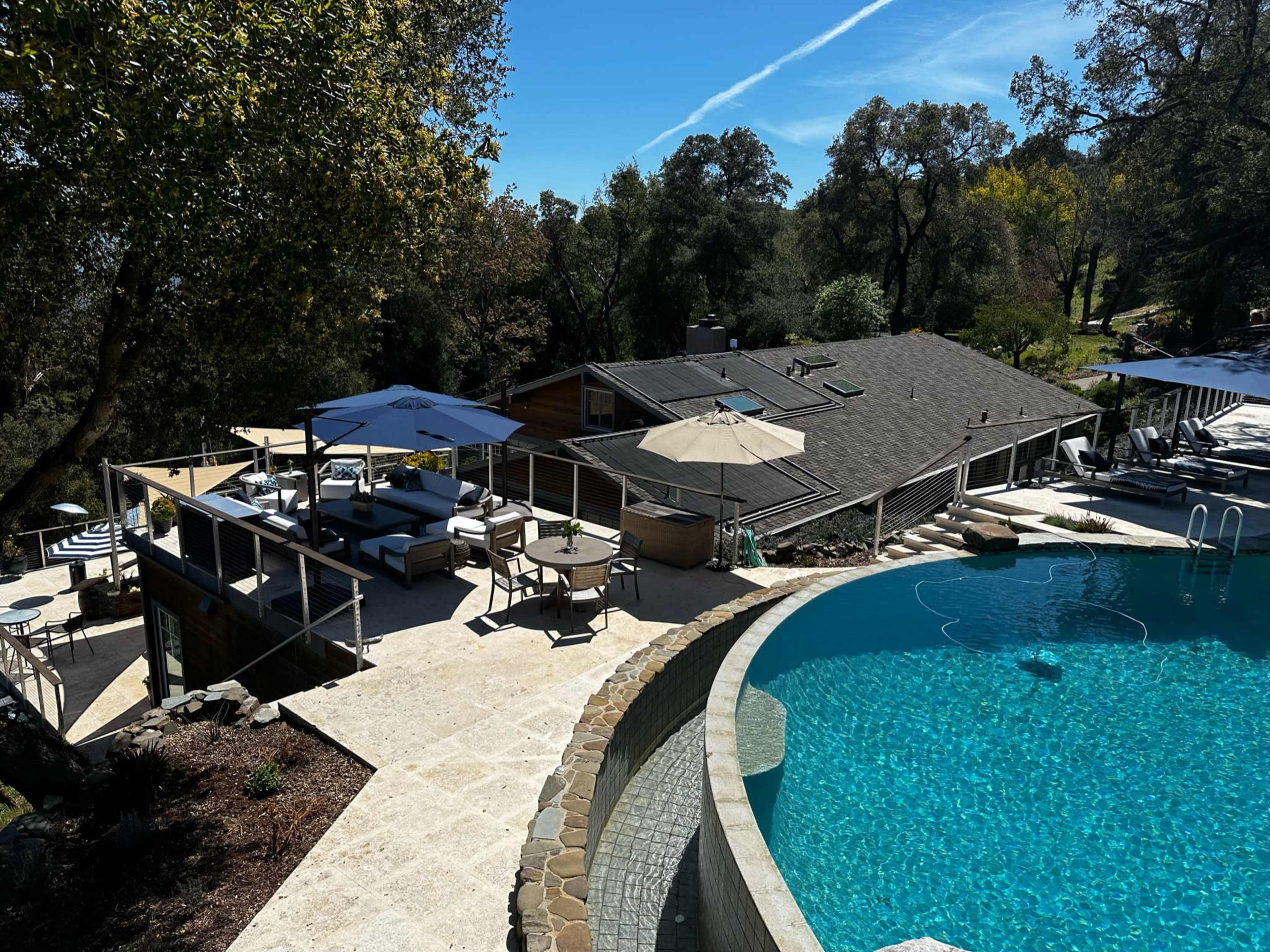 A house with a large swimming pool and a patio area, surrounded by trees and outdoor seating.