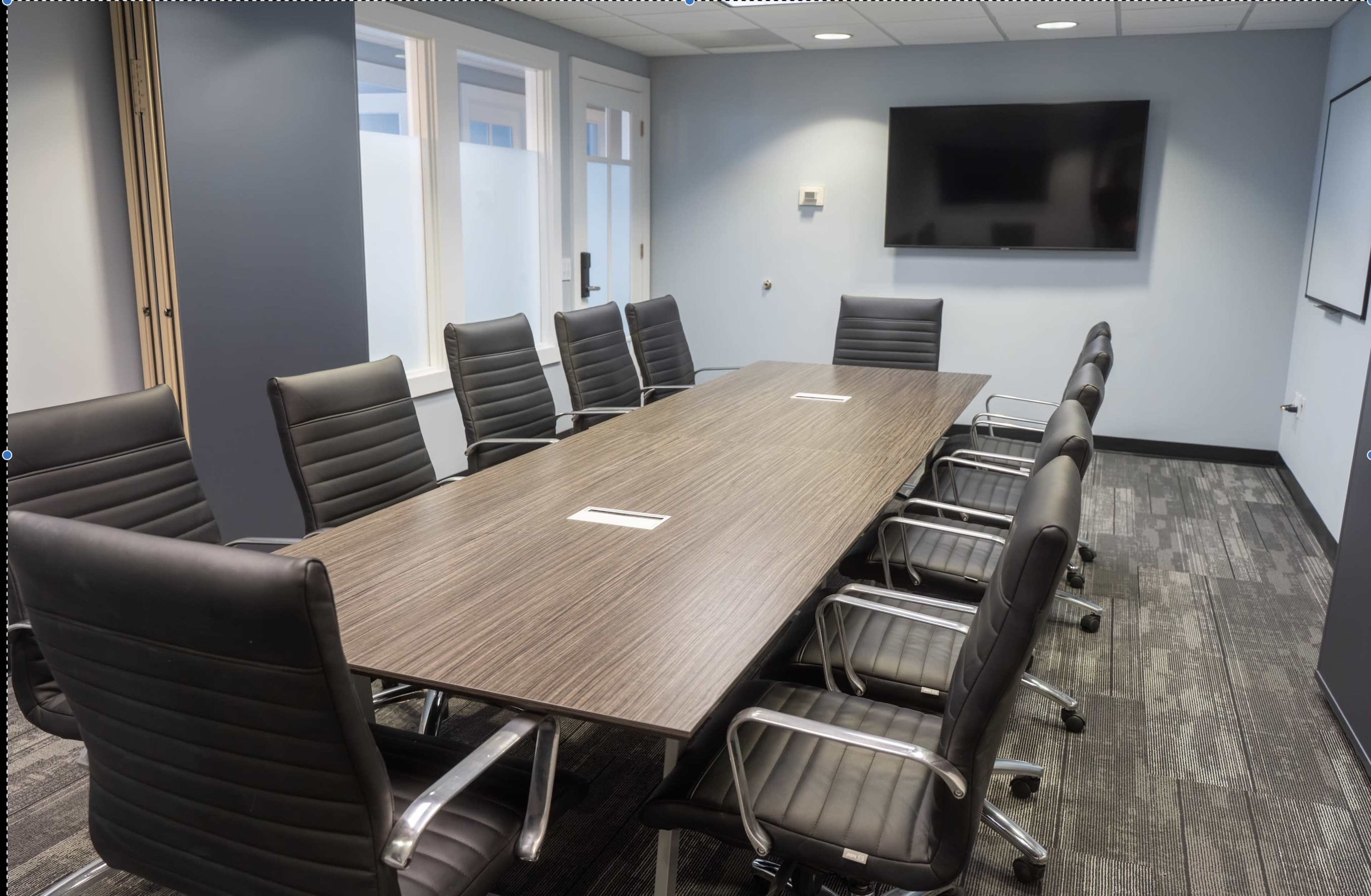 A modern conference room features a long wooden table surrounded by ergonomic black chairs, with a large screen mounted on the wall.
