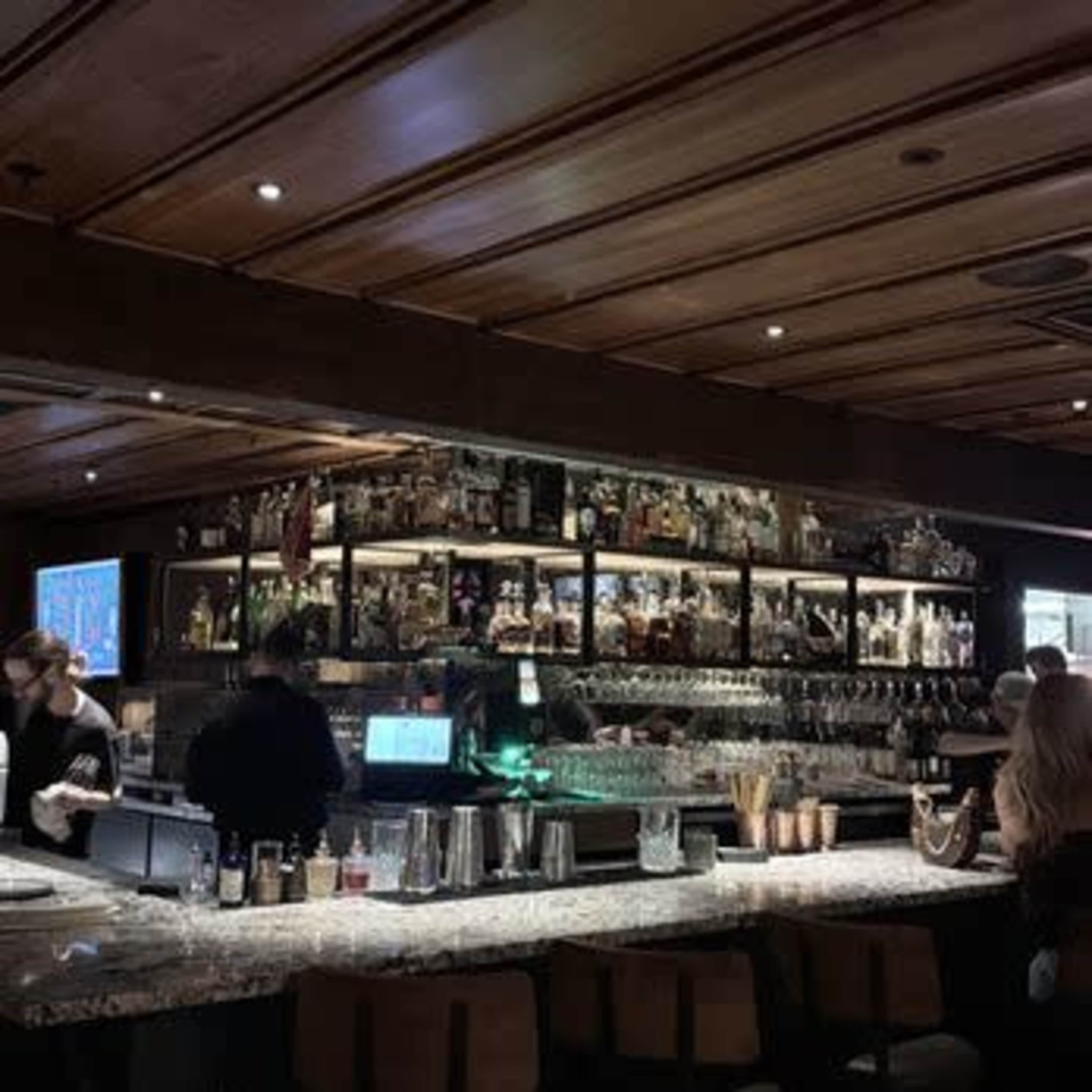 The image shows a bar area with shelves lined with various bottles, a marble countertop, and patrons seated along the bar.