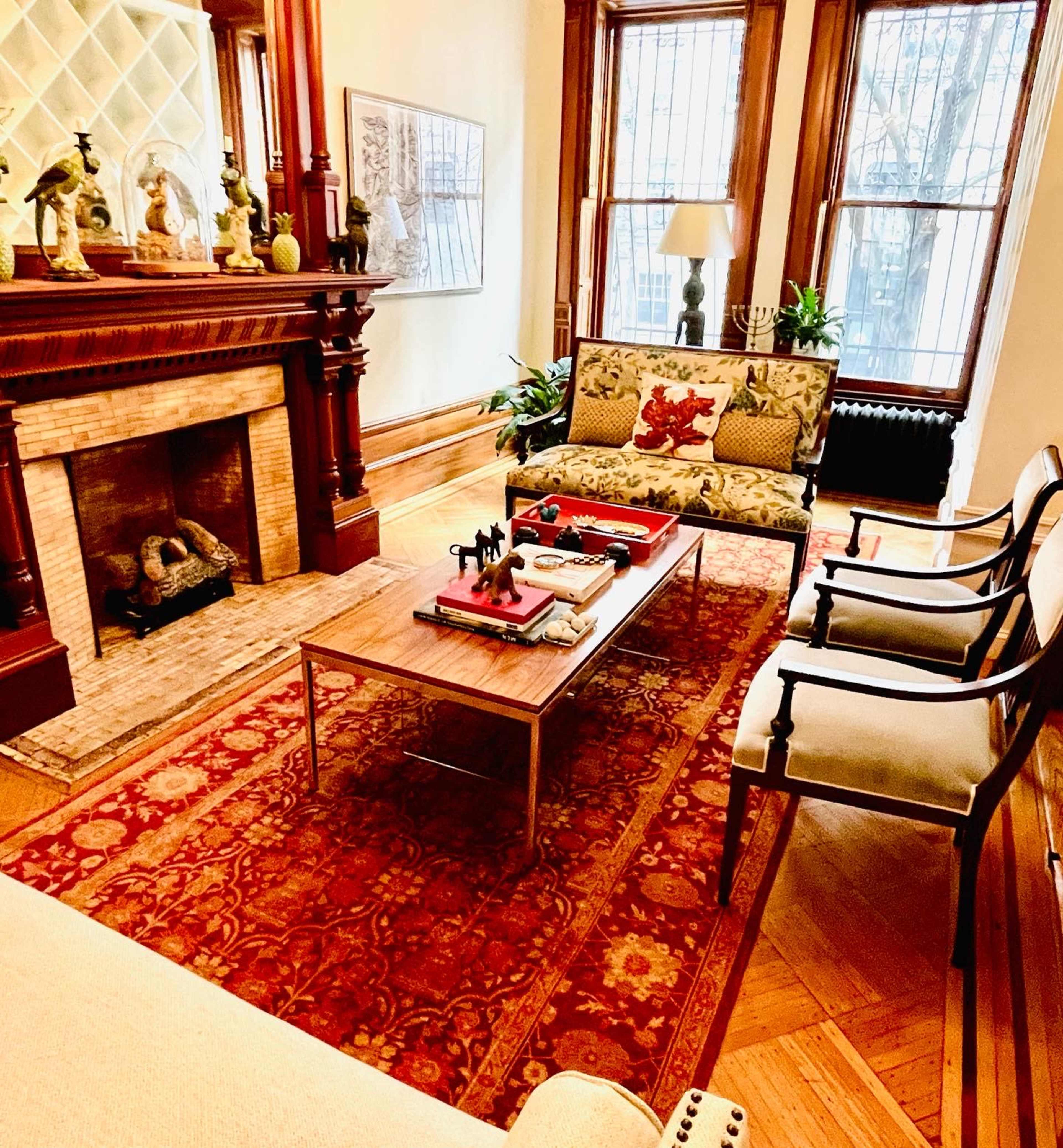 A cozy living room with a fireplace, two upholstered chairs, a coffee table adorned with decor, and large windows allowing natural light.