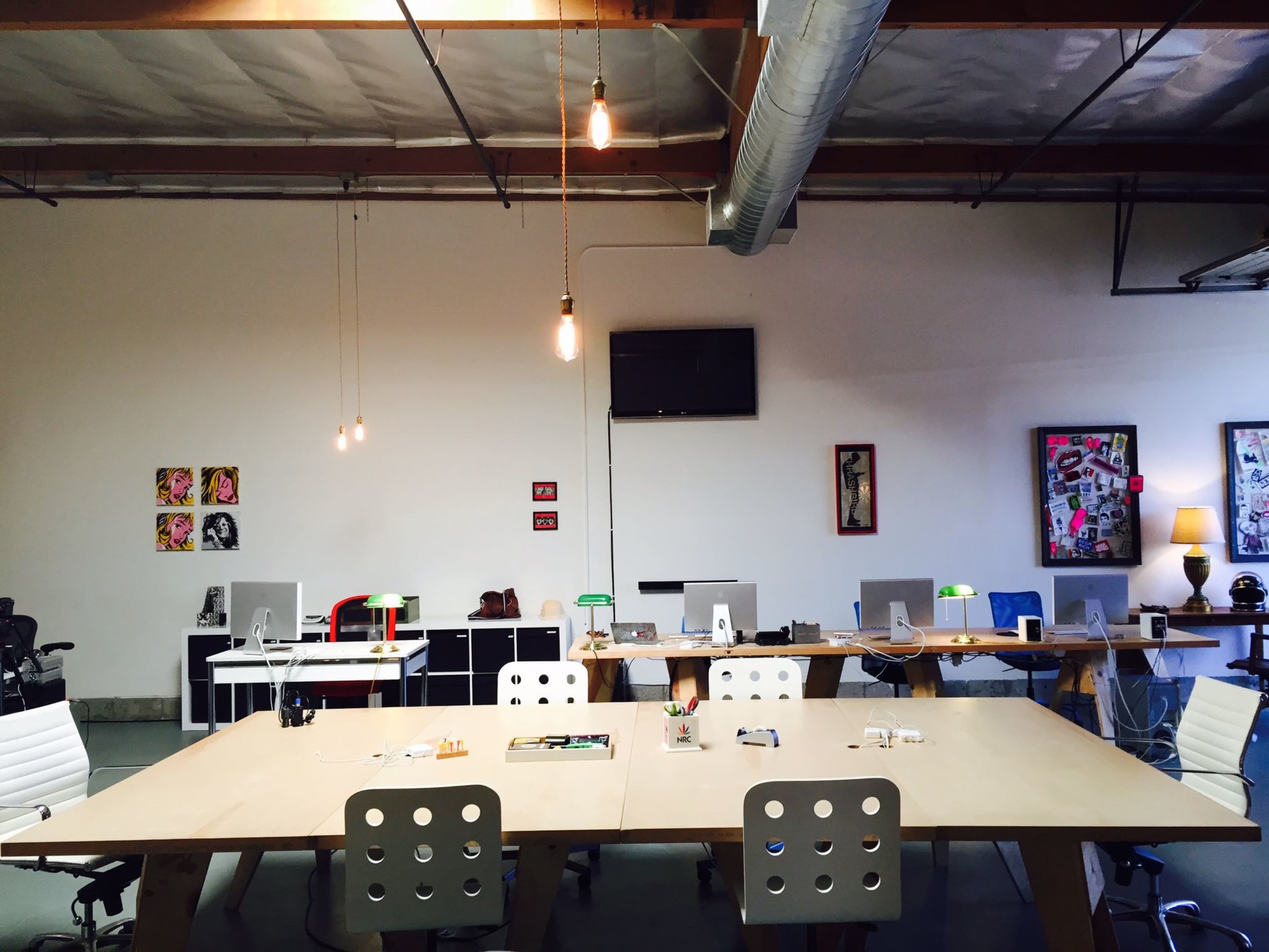 An office space features multiple desks with computers, hanging lights, and decorative wall art.