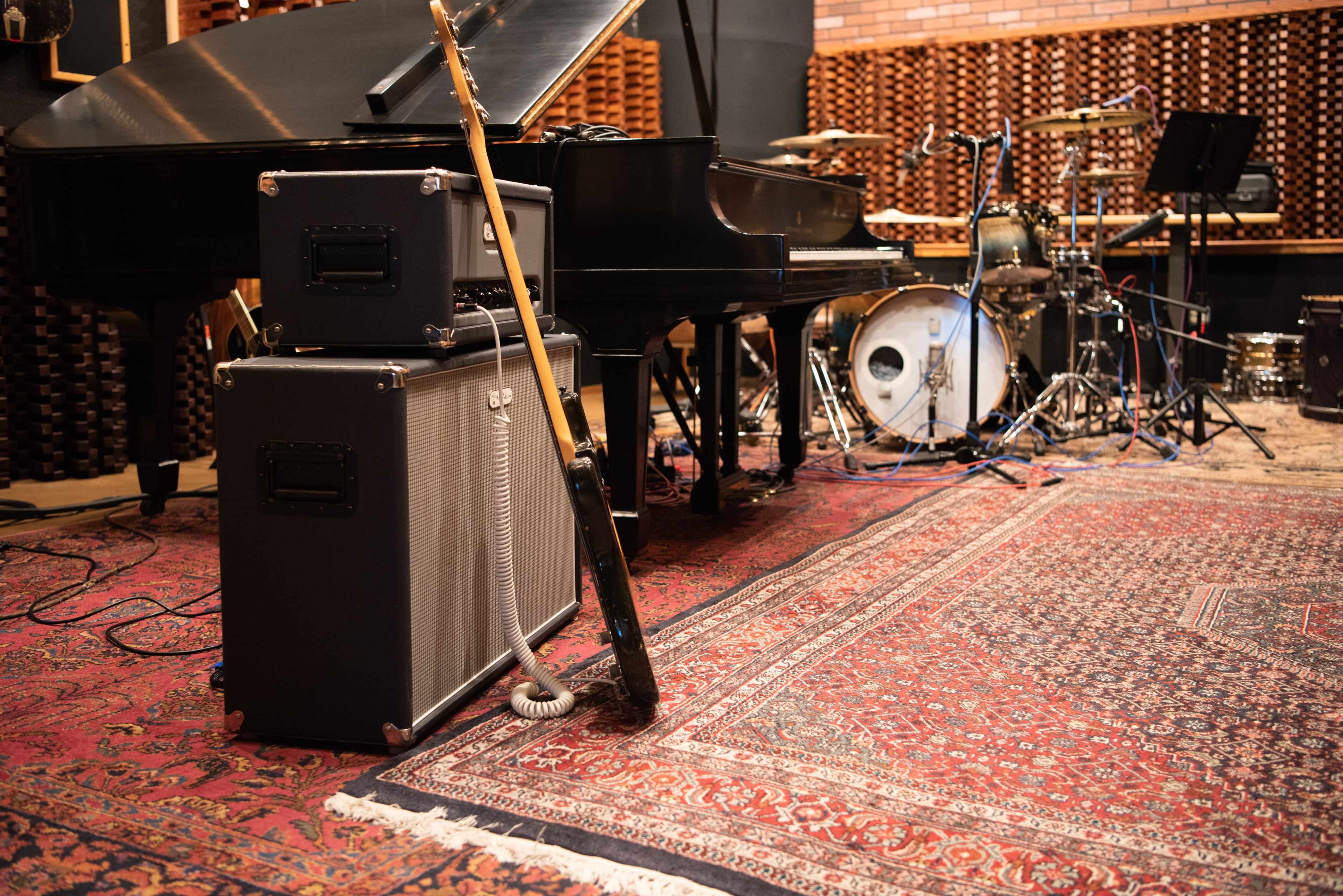 A music studio featuring a grand piano, amplifiers, a guitar, and a drum set arranged on a patterned rug.