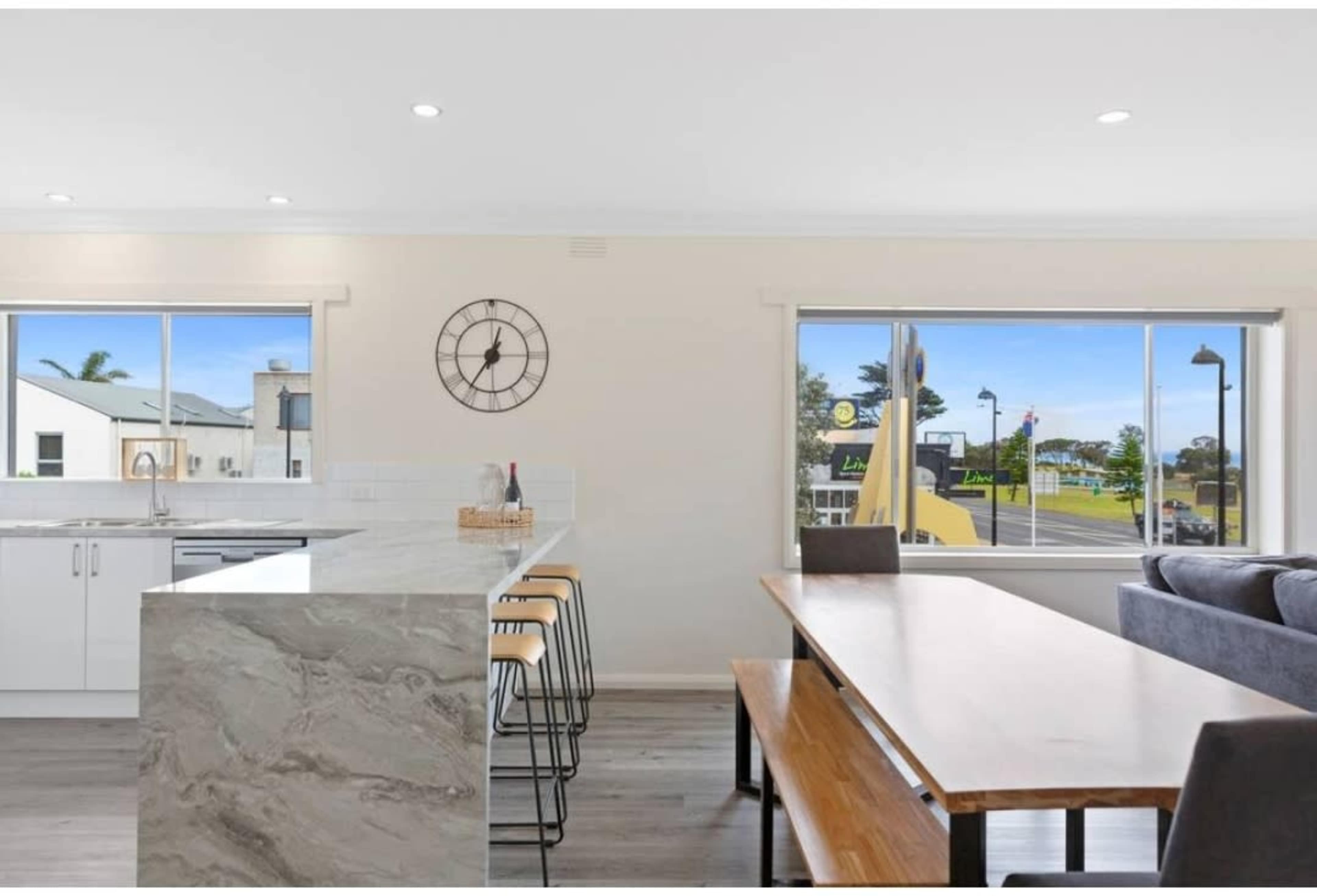 A modern kitchen and dining area features a large island with bar stools and a wooden table by a window overlooking a street.