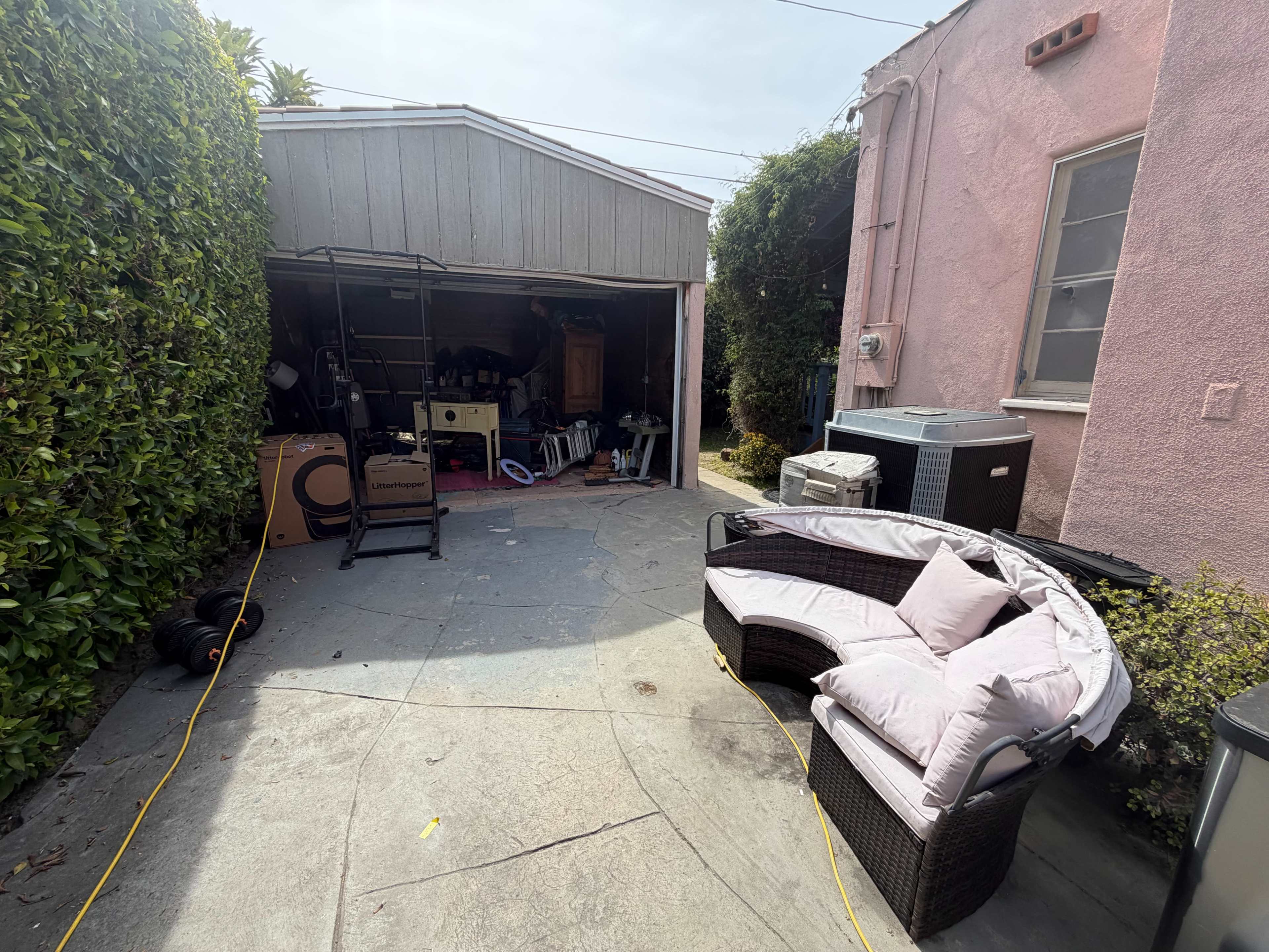 The image shows a small outdoor space with a curved wicker sofa in front of a garage filled with various items, surrounded by greenery and a pink building.