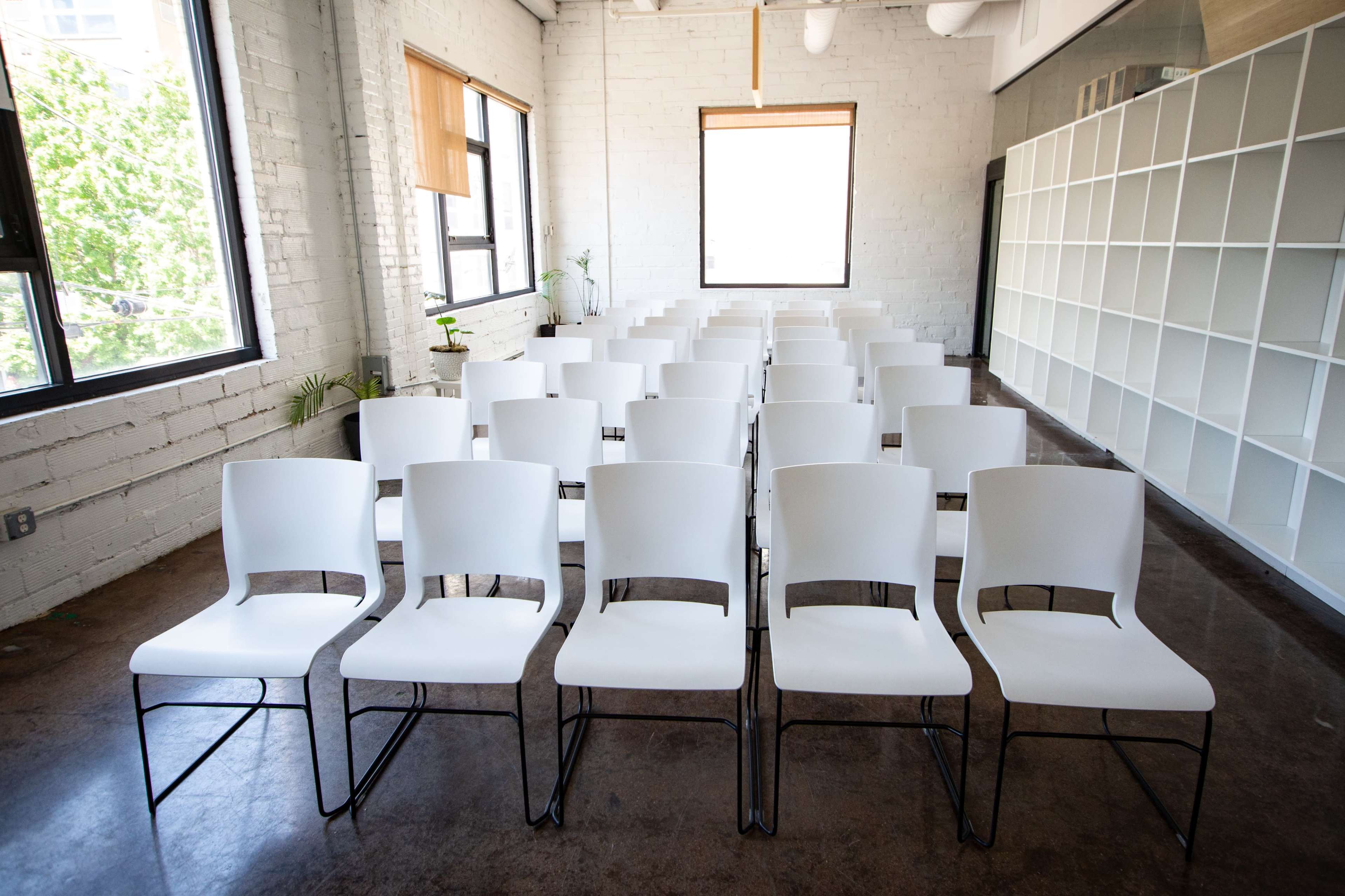 The image shows a neatly arranged row of white chairs in a bright, modern room with large windows.