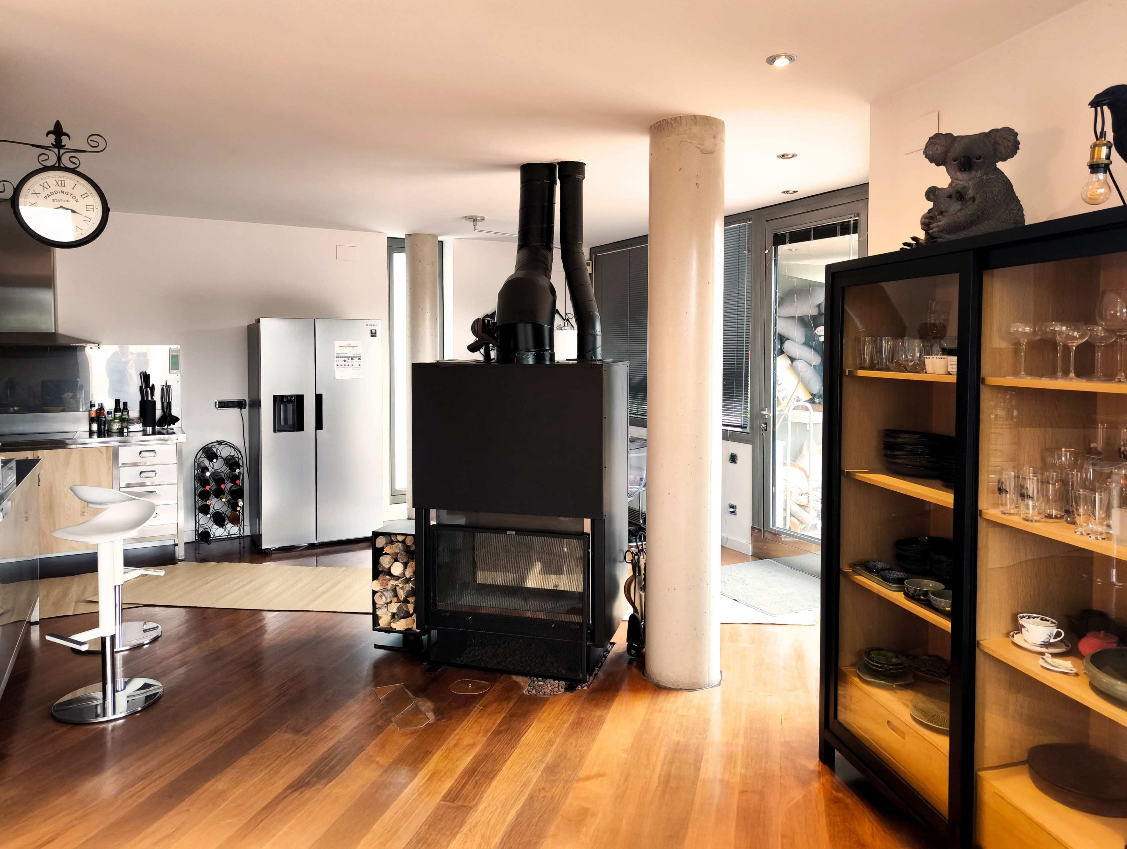 The image shows a modern kitchen and living area featuring a black fireplace, a white refrigerator, and a glass cabinet displaying dishware.