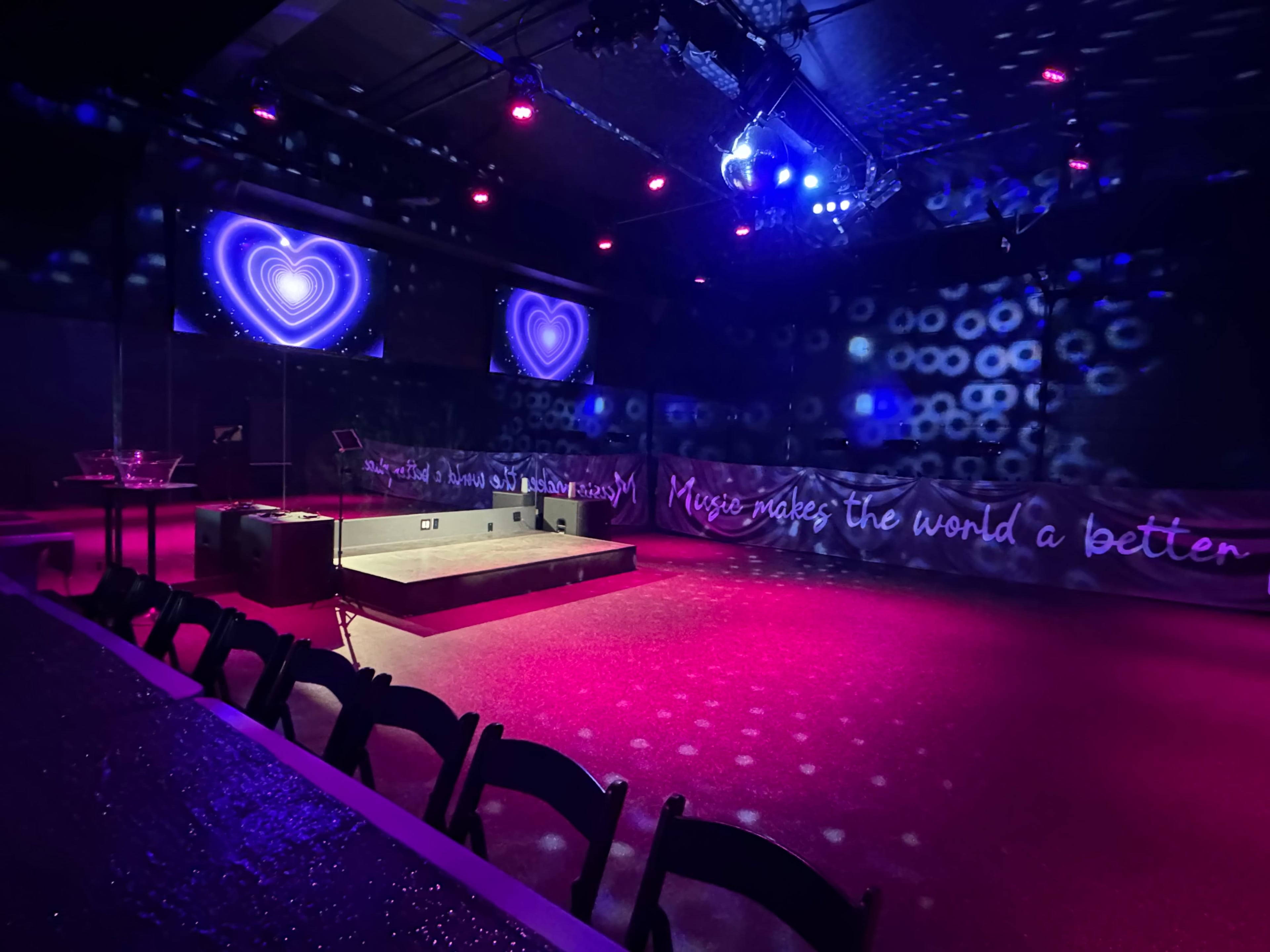 The image shows a dimly lit music venue featuring a stage with bright lights, heart-shaped visuals on screens, and a wall decorated with a quote about music.
