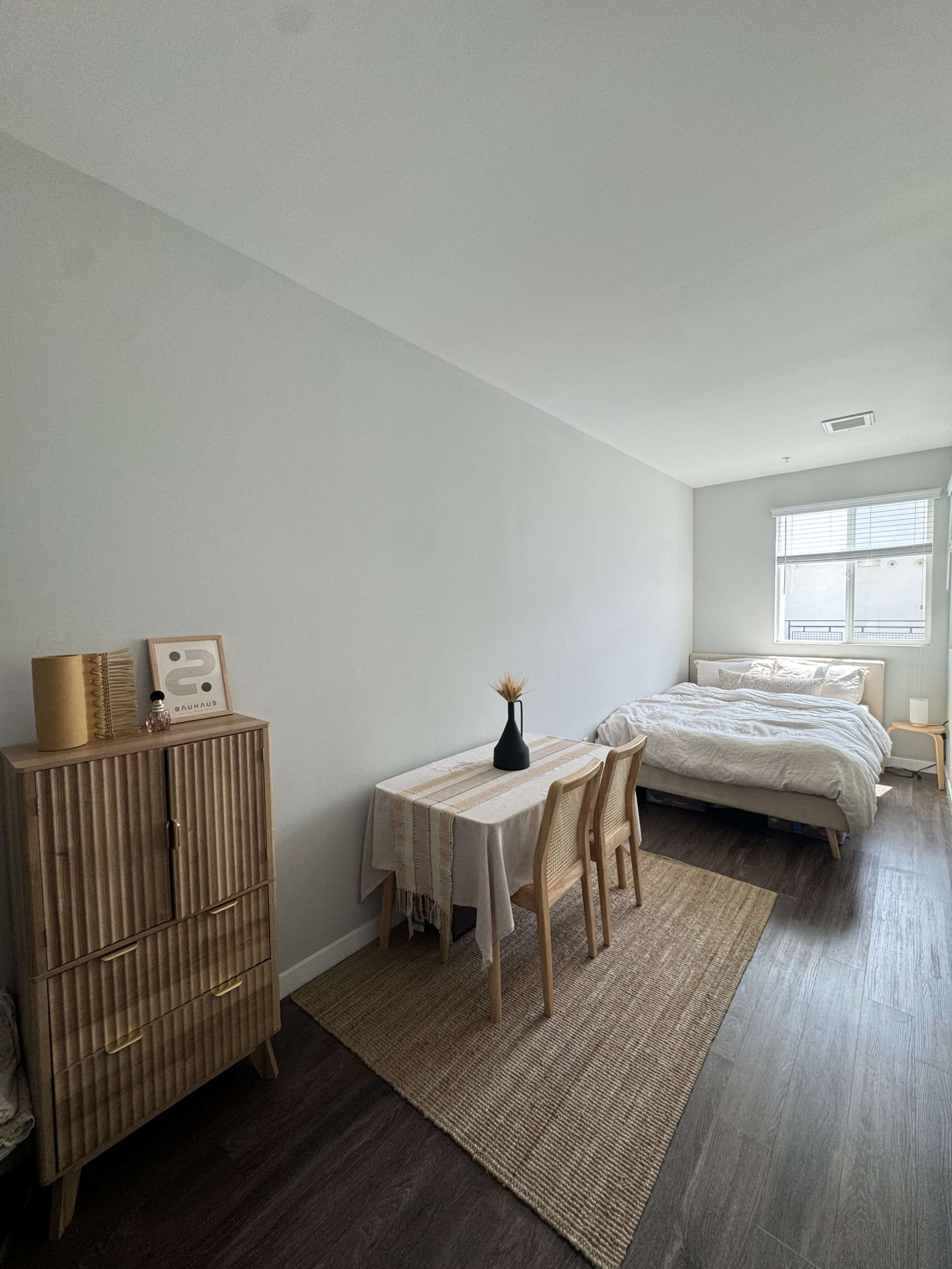 The image shows a minimalist bedroom featuring a bed, a small dining table with chairs, a vase, and a wooden storage unit against a light-colored wall.
