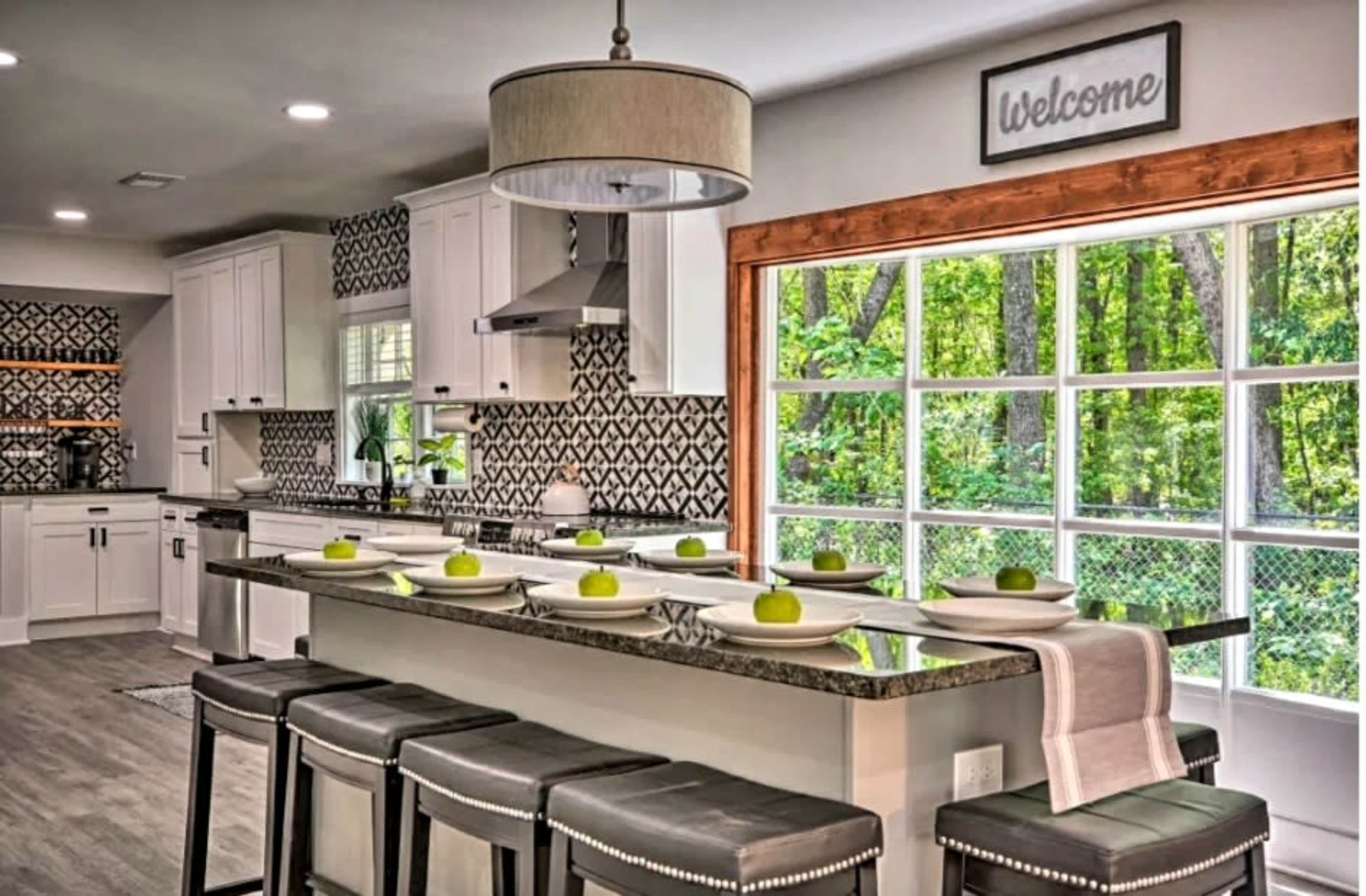 The kitchen features a modern design with white cabinetry, a large island topped with green apples, and a window overlooking a wooded area.