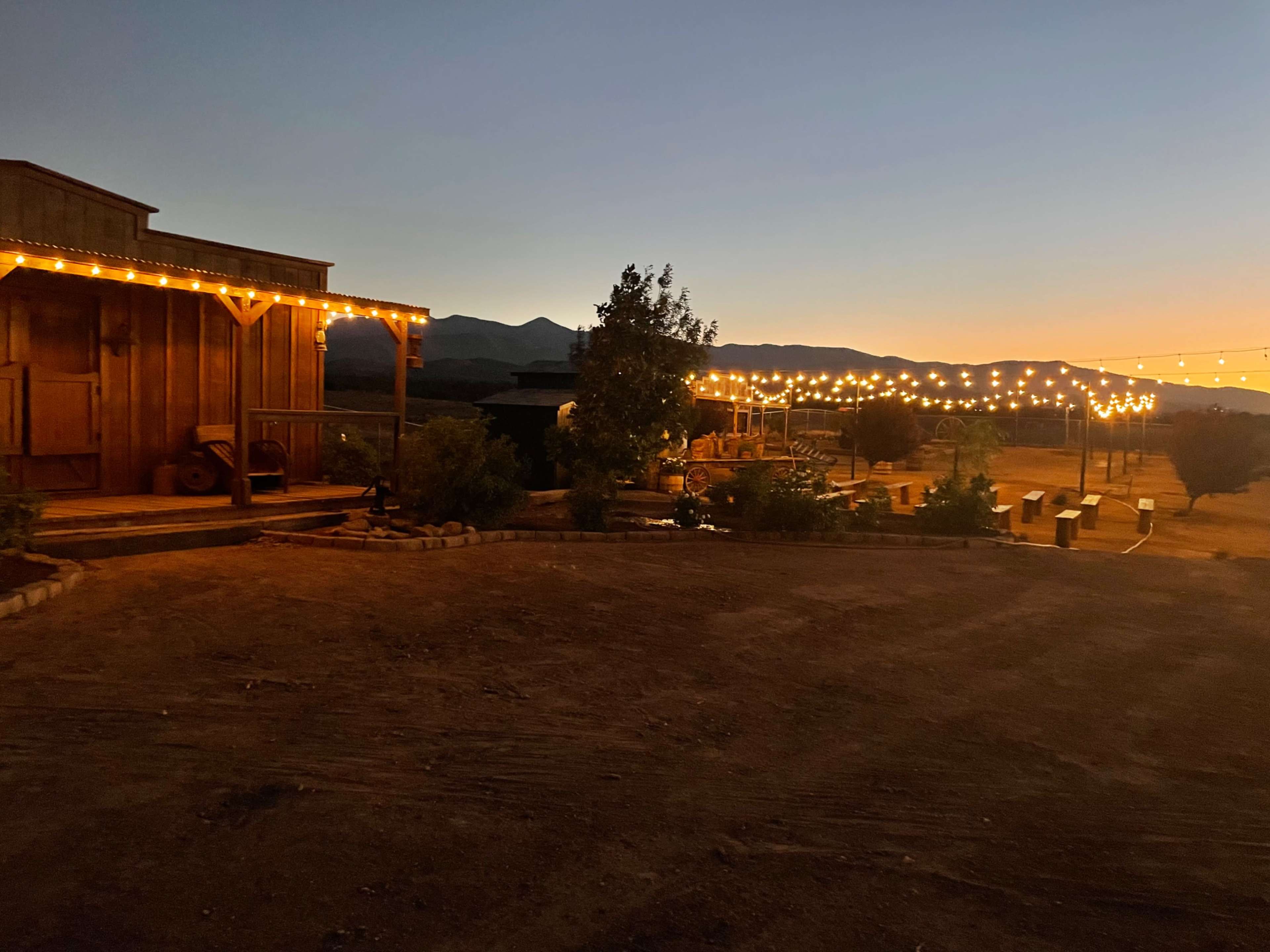 A rustic wooden building illuminated by string lights at dusk, with mountains in the background and a dirt path leading to additional structures.