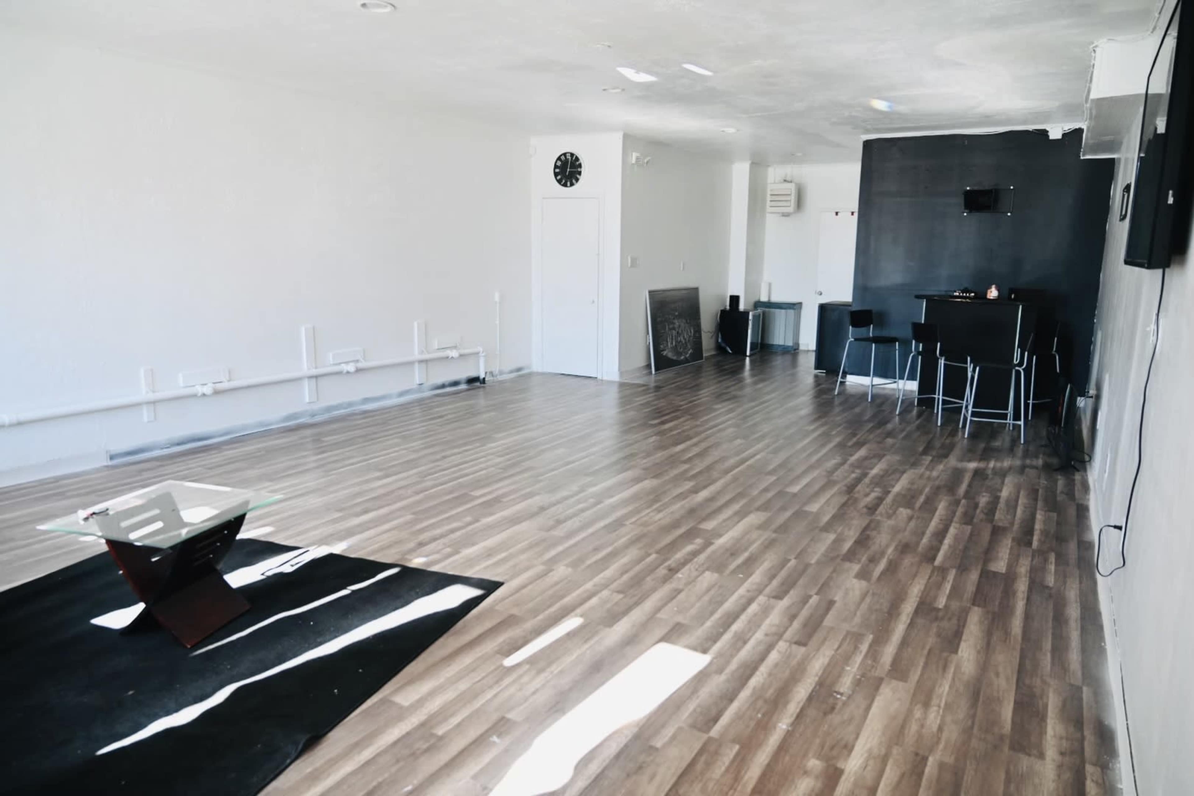 A spacious, empty room with wooden flooring, a black feature wall, a clock on one wall, and minimal furnishings, including a glass table and bar stools.