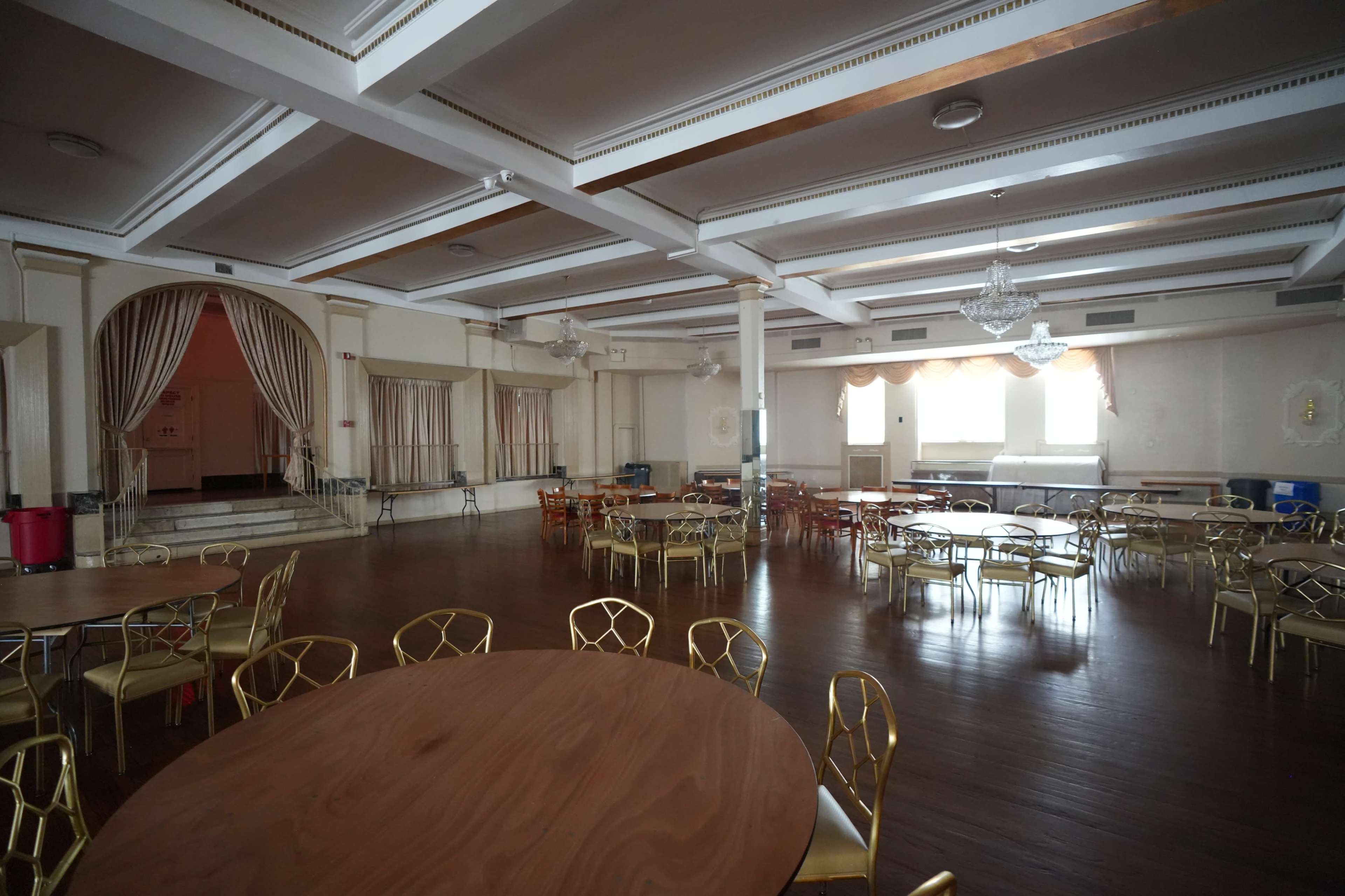 The image shows a large, empty event hall with round tables and chairs arranged throughout the space, featuring a stage and decorative lighting.