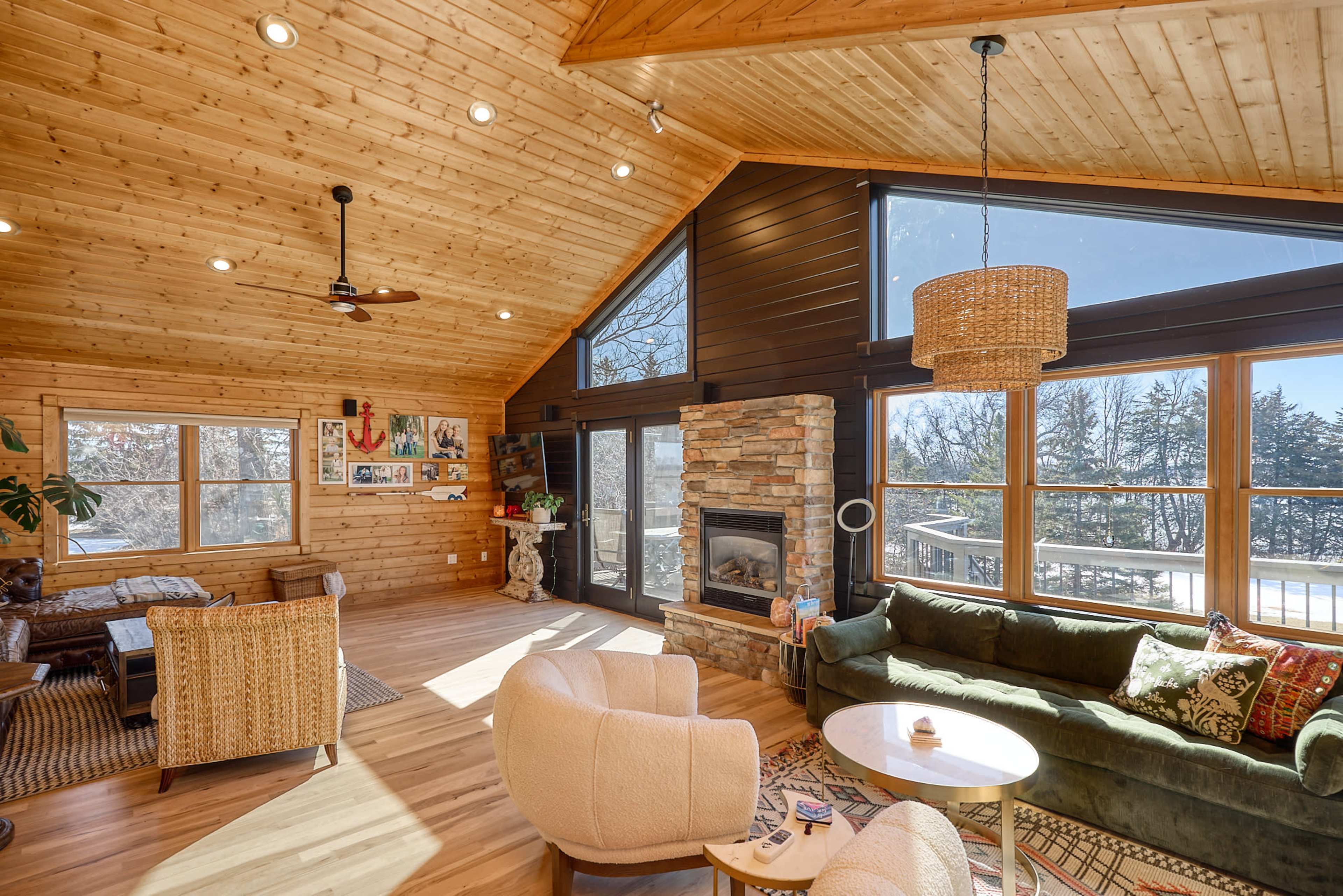The image shows a spacious wooden cabin interior with large windows, a stone fireplace, and cozy seating areas arranged around a coffee table.