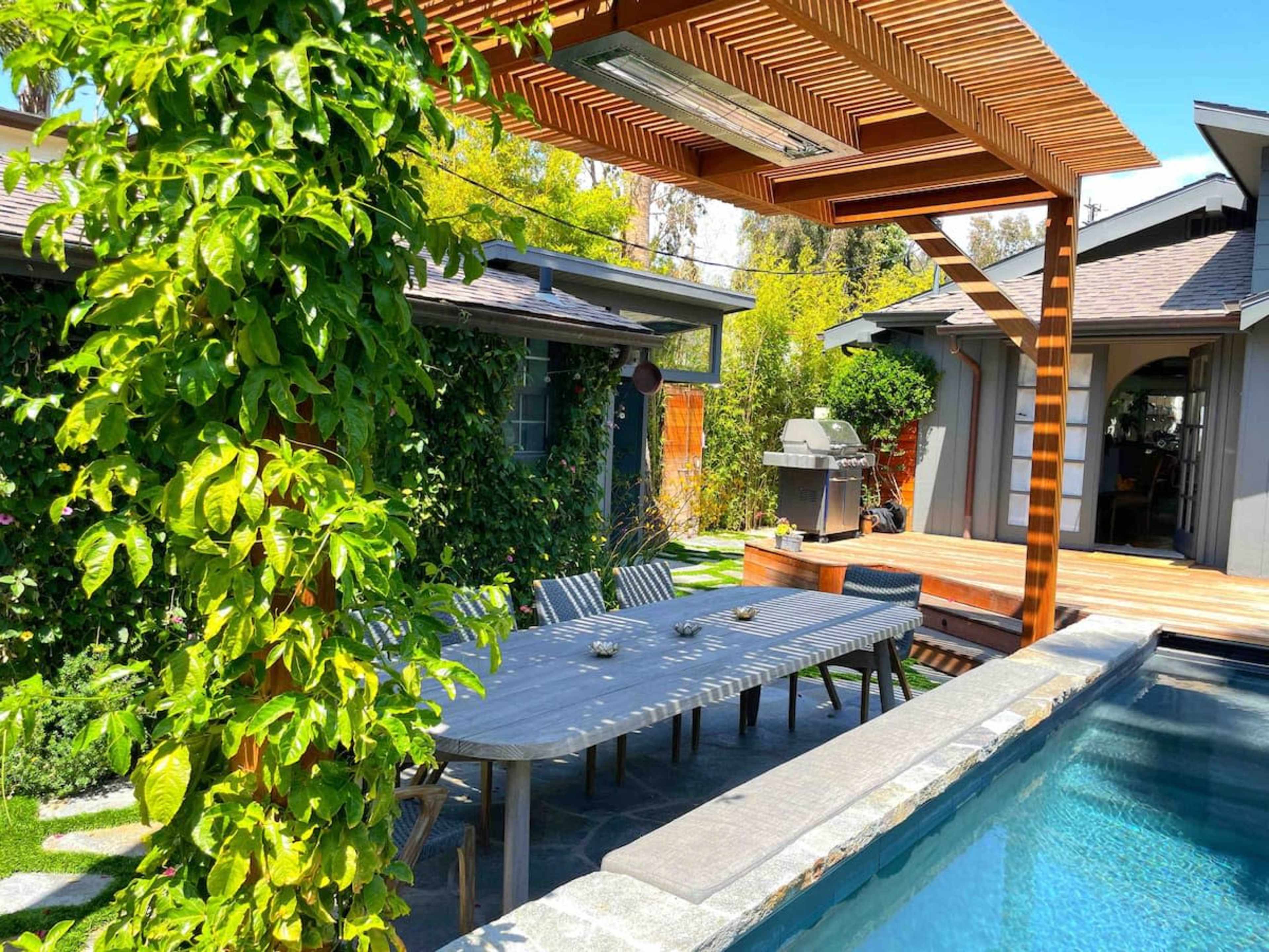 A wooden pergola shades a dining table beside a pool in a landscaped backyard with a grill and lush greenery.