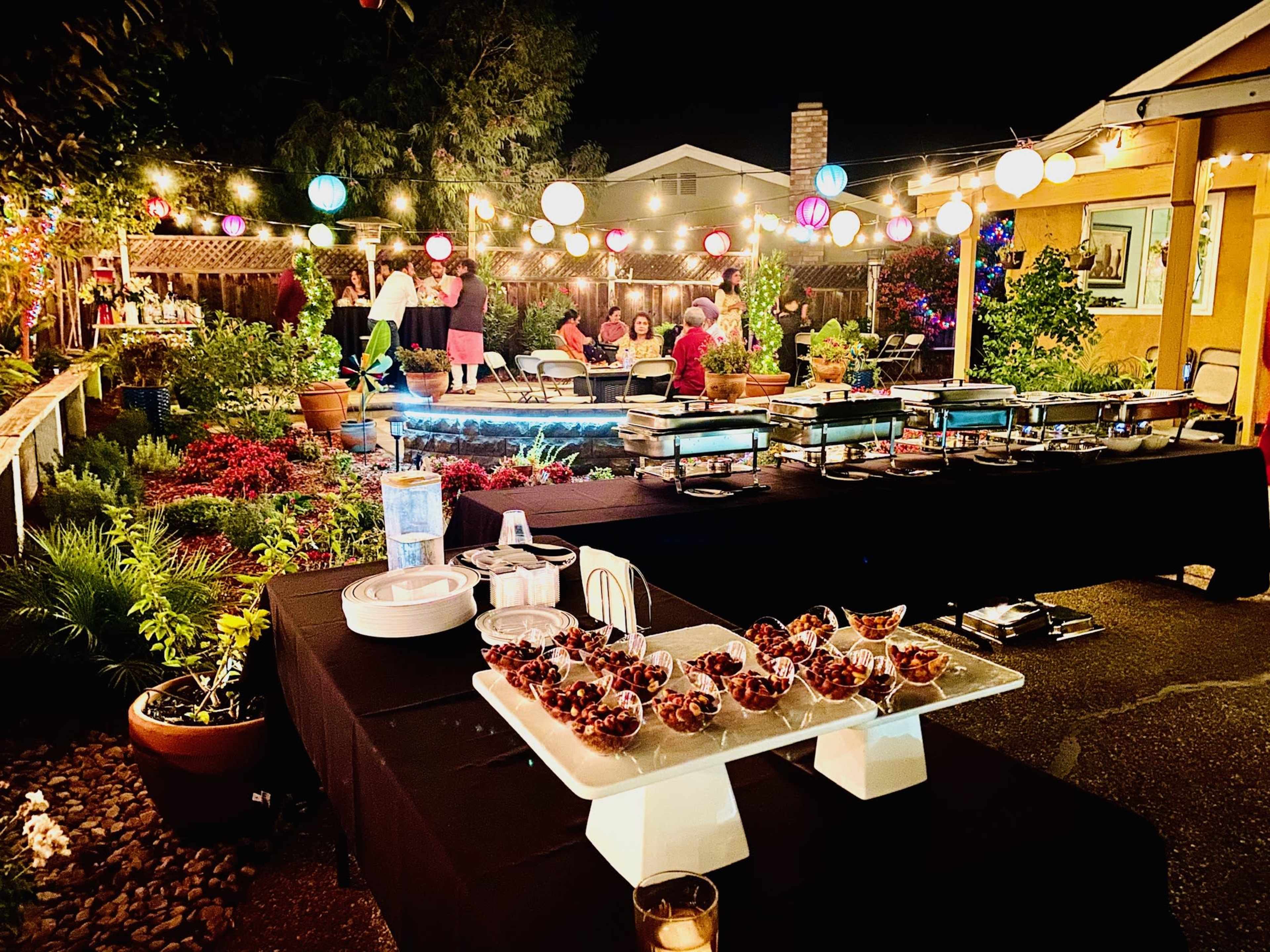 The scene depicts an outdoor evening gathering with tables of food and colorful lanterns illuminating the garden setting.
