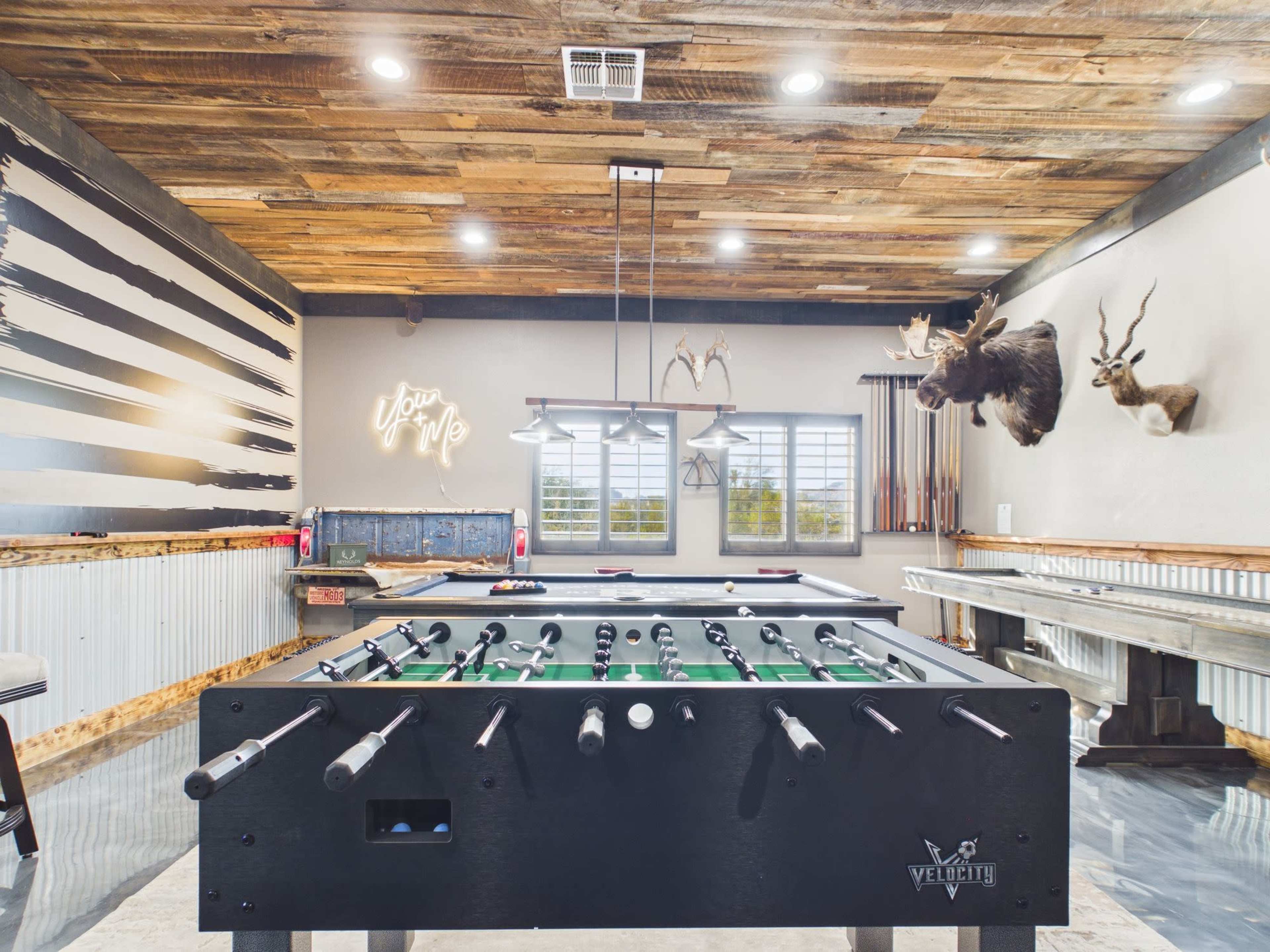 The image depicts a recreational room featuring a foosball table, wooden walls with mounted animal heads, and lighting fixtures above.