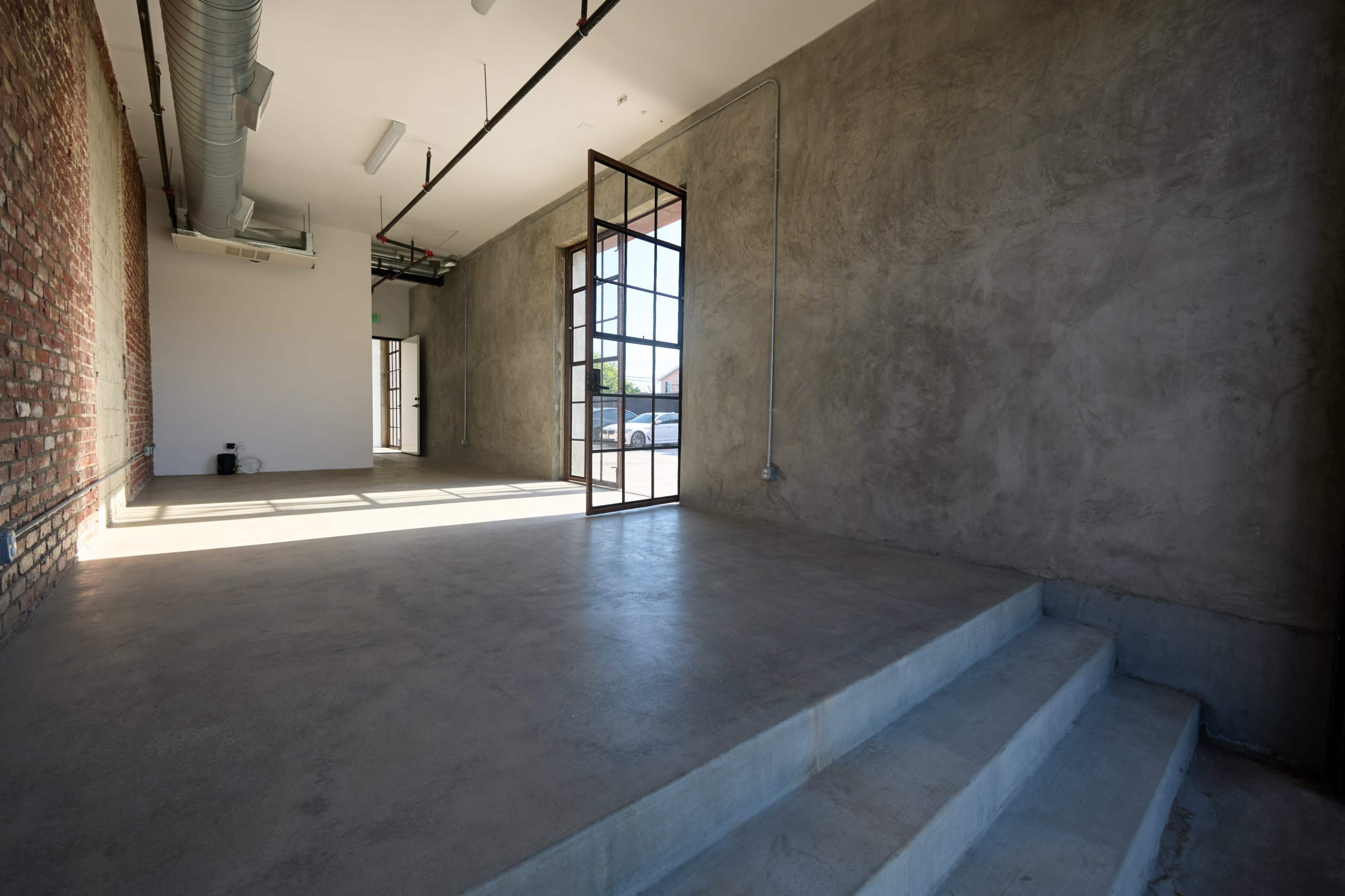 The image shows an empty interior space with a concrete floor, a mix of exposed brick and smooth concrete walls, and large glass doors leading outside.