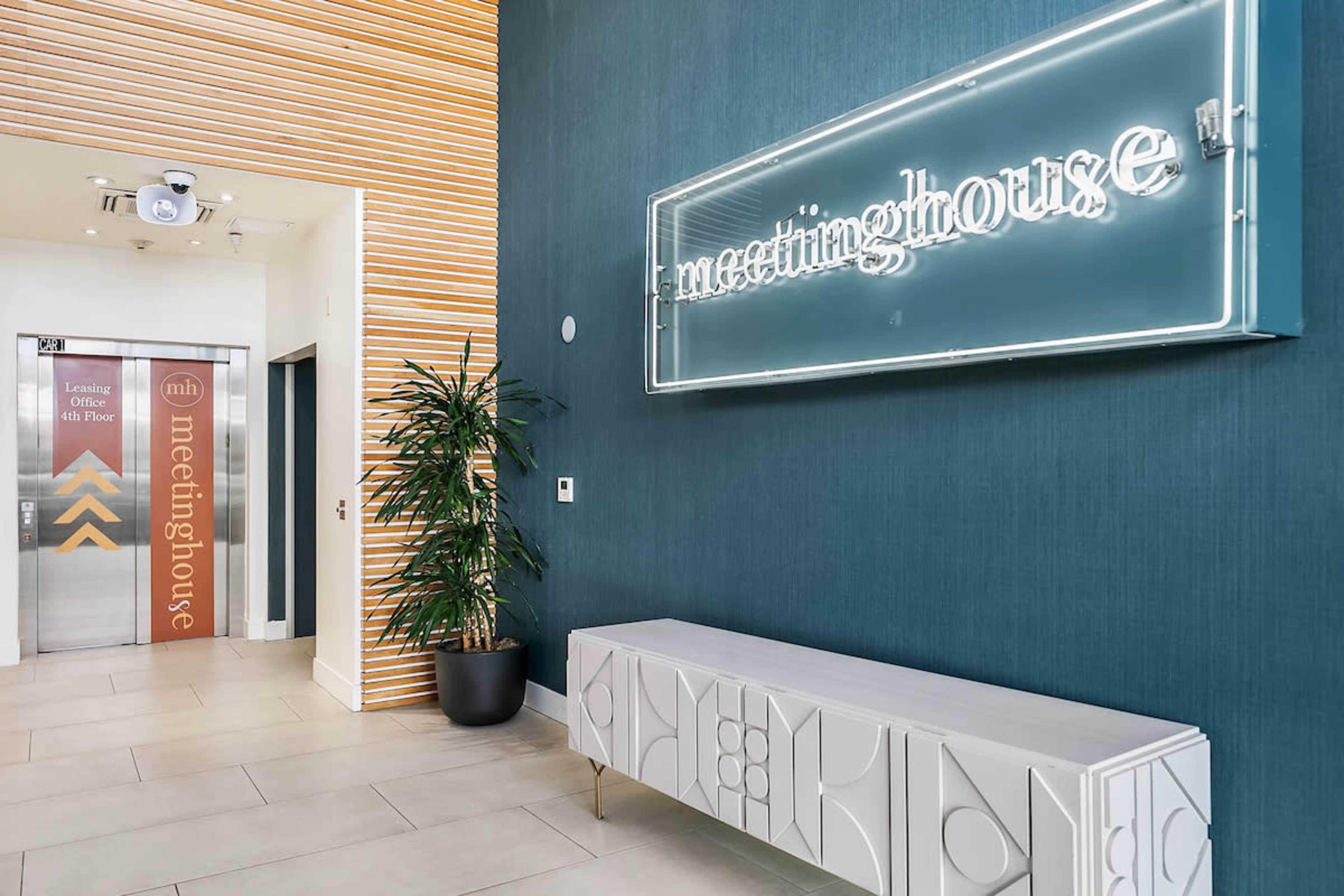 The image shows a modern lobby featuring a neon sign that says "meetinghouse," a decorative bench, and an elevator with a directional sign indicating various floors.