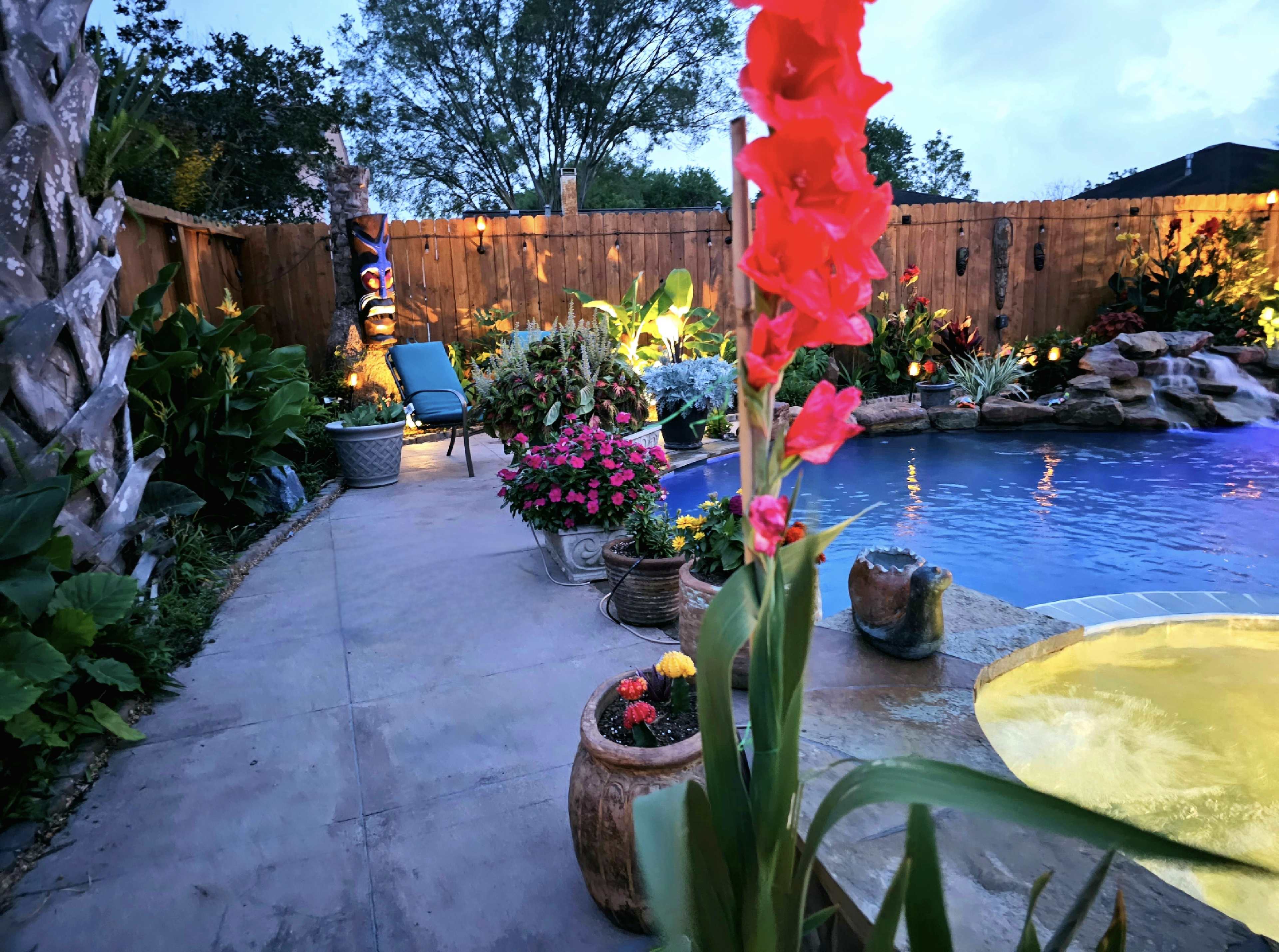 A landscaped backyard featuring a swimming pool surrounded by colorful flowers and plants, with a seating area and decorative accents.