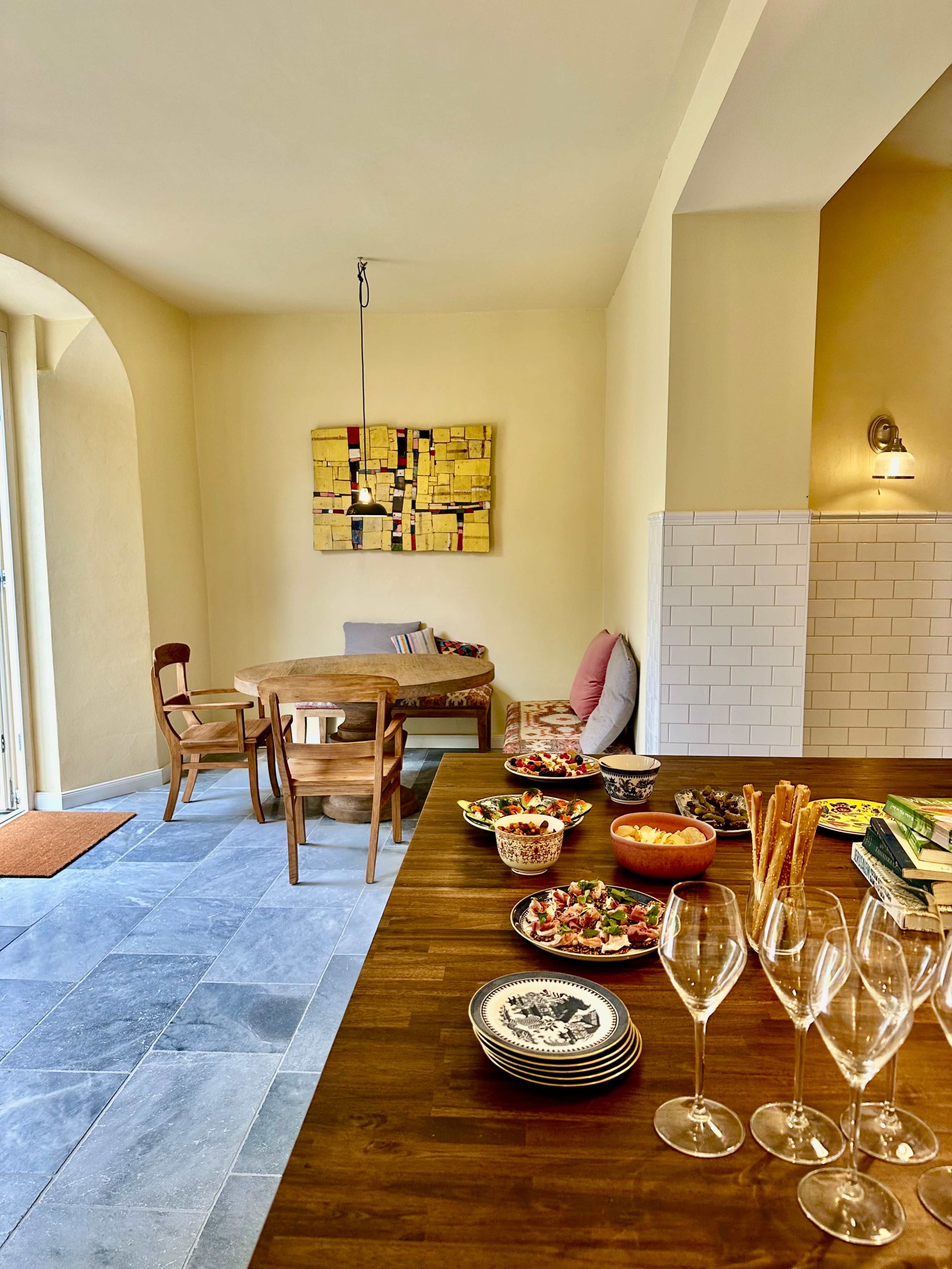 The image shows a brightly lit dining area with a wooden table set with various dishes and glasses, a beige wall with an abstract painting, and a cozy seating arrangement in the background.