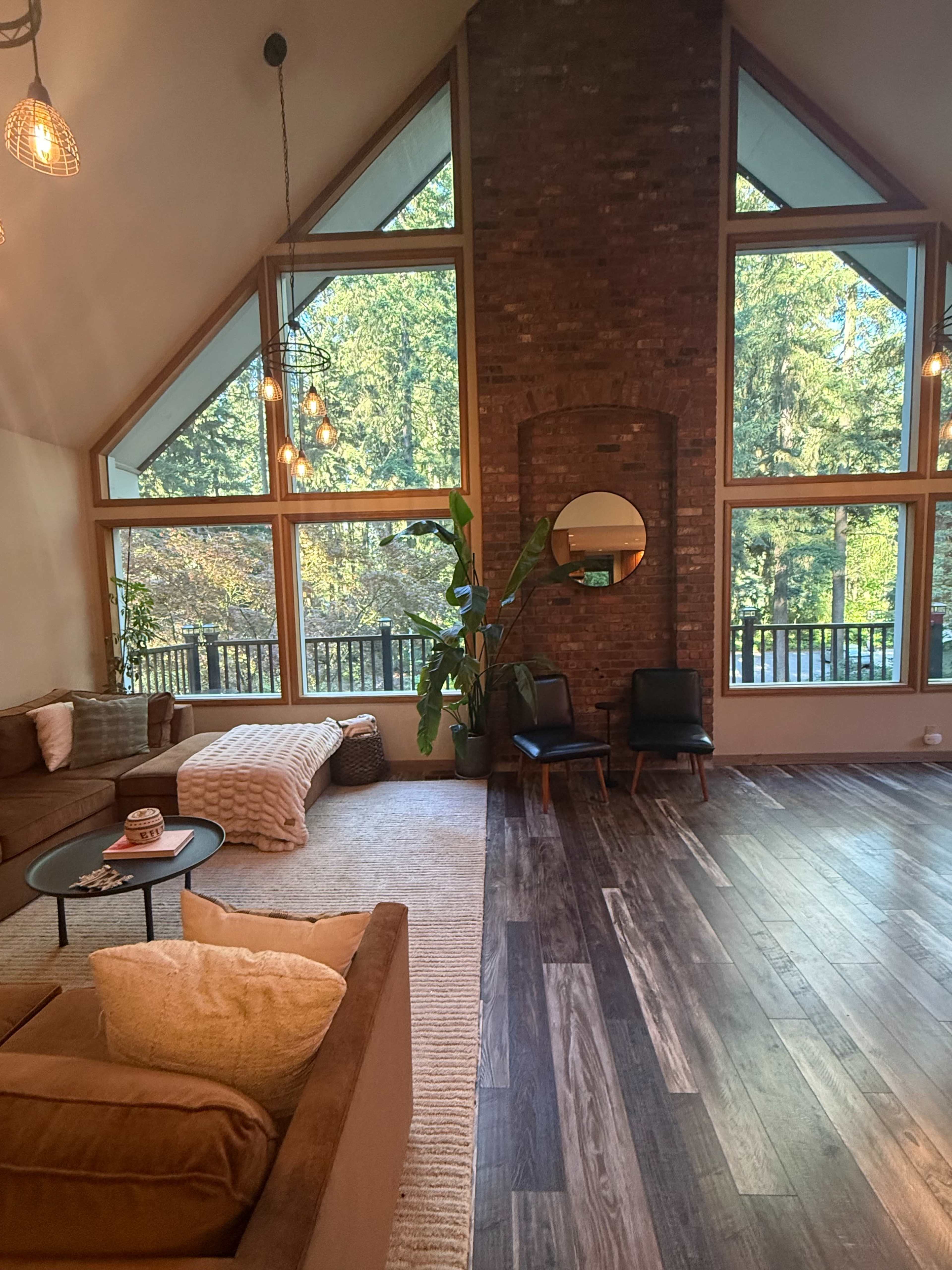 The spacious living room features large windows overlooking a forest, with a brick wall, a round mirror, modern seating, and a cozy area rug.