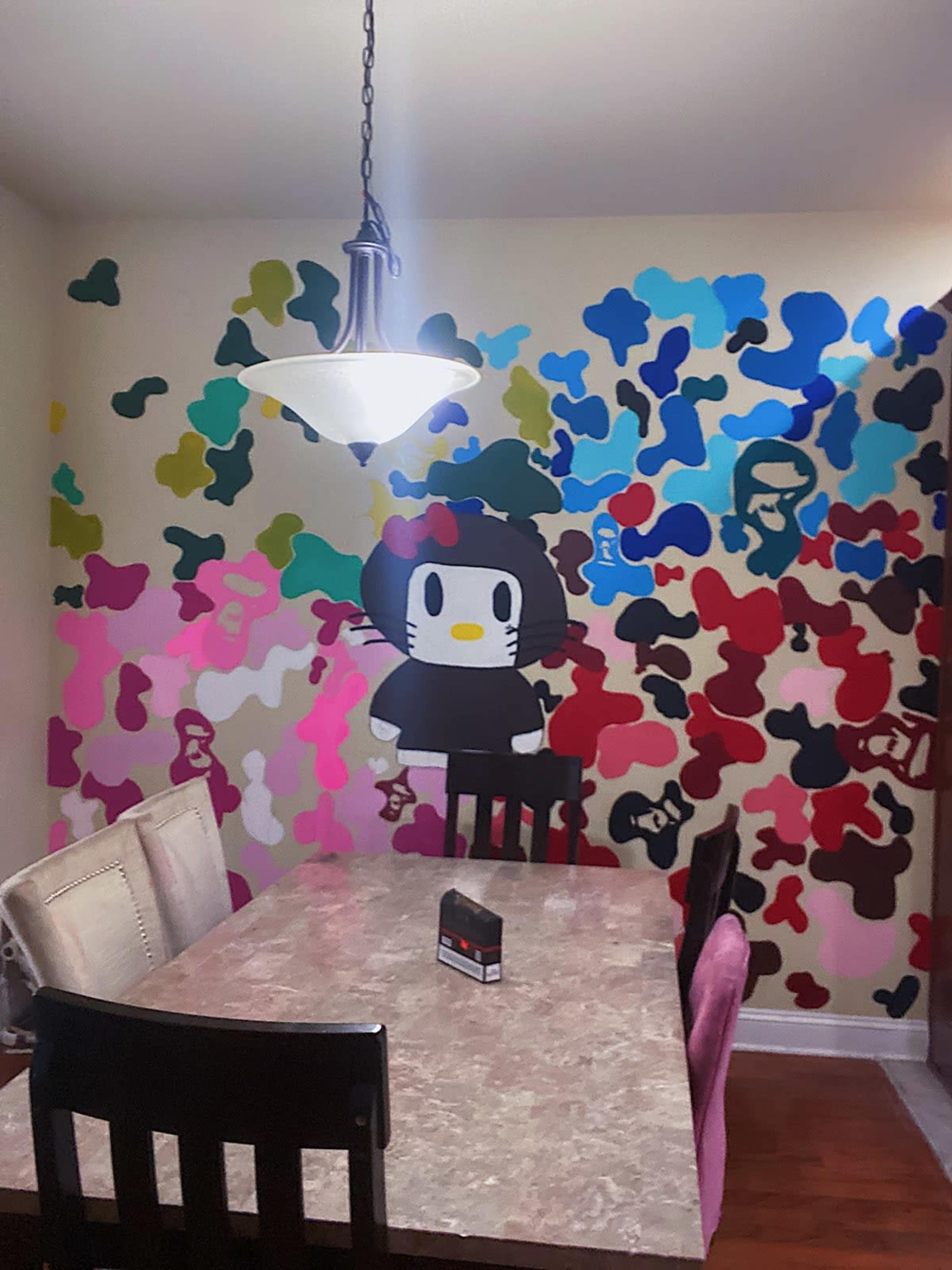 The image shows a dining area with a table and chairs, featuring a colorful wall mural of cartoon characters and abstract shapes.