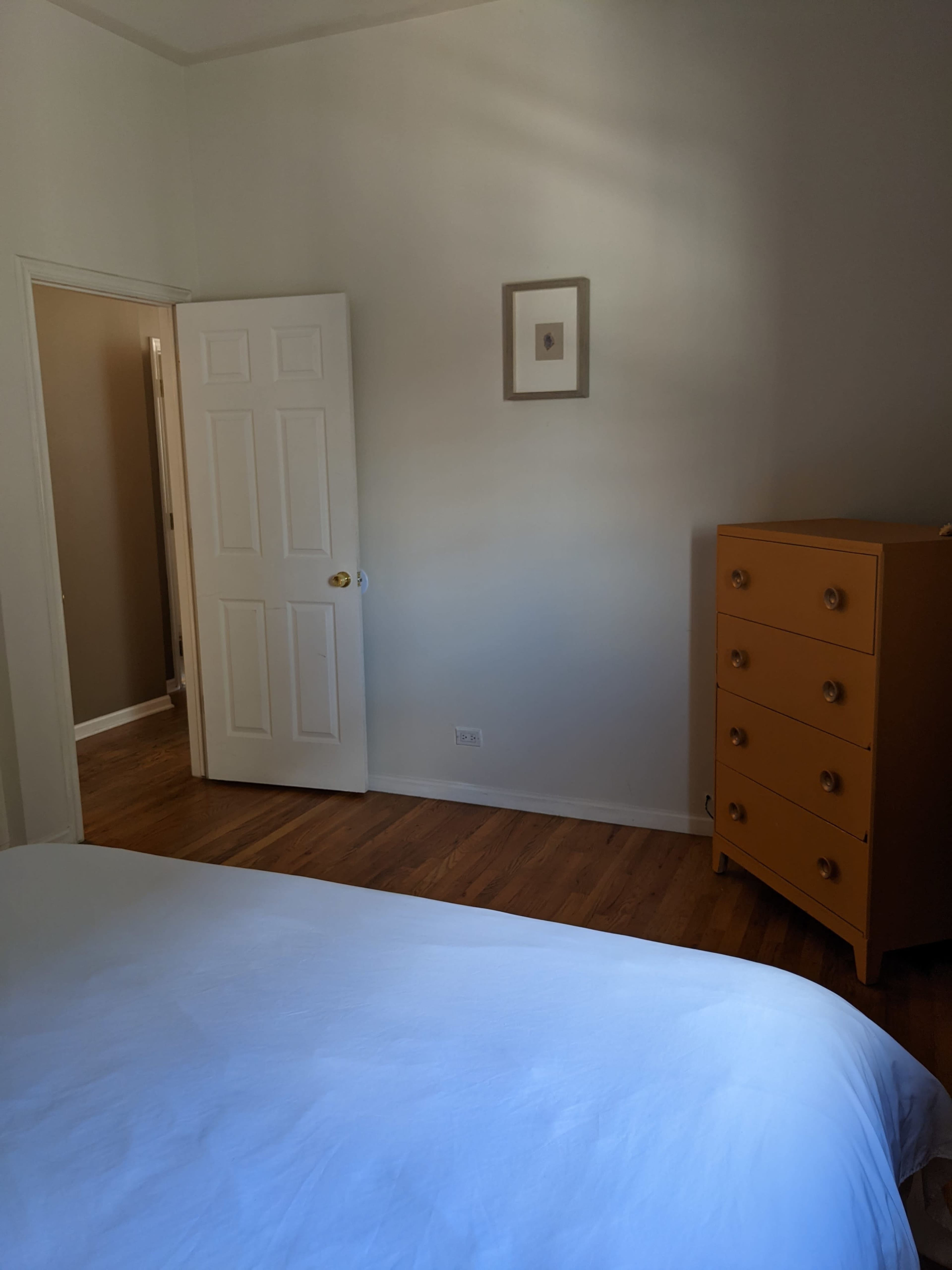 A white bed is positioned in a well-lit bedroom with an open door leading to another room and a tall wooden dresser in the corner.