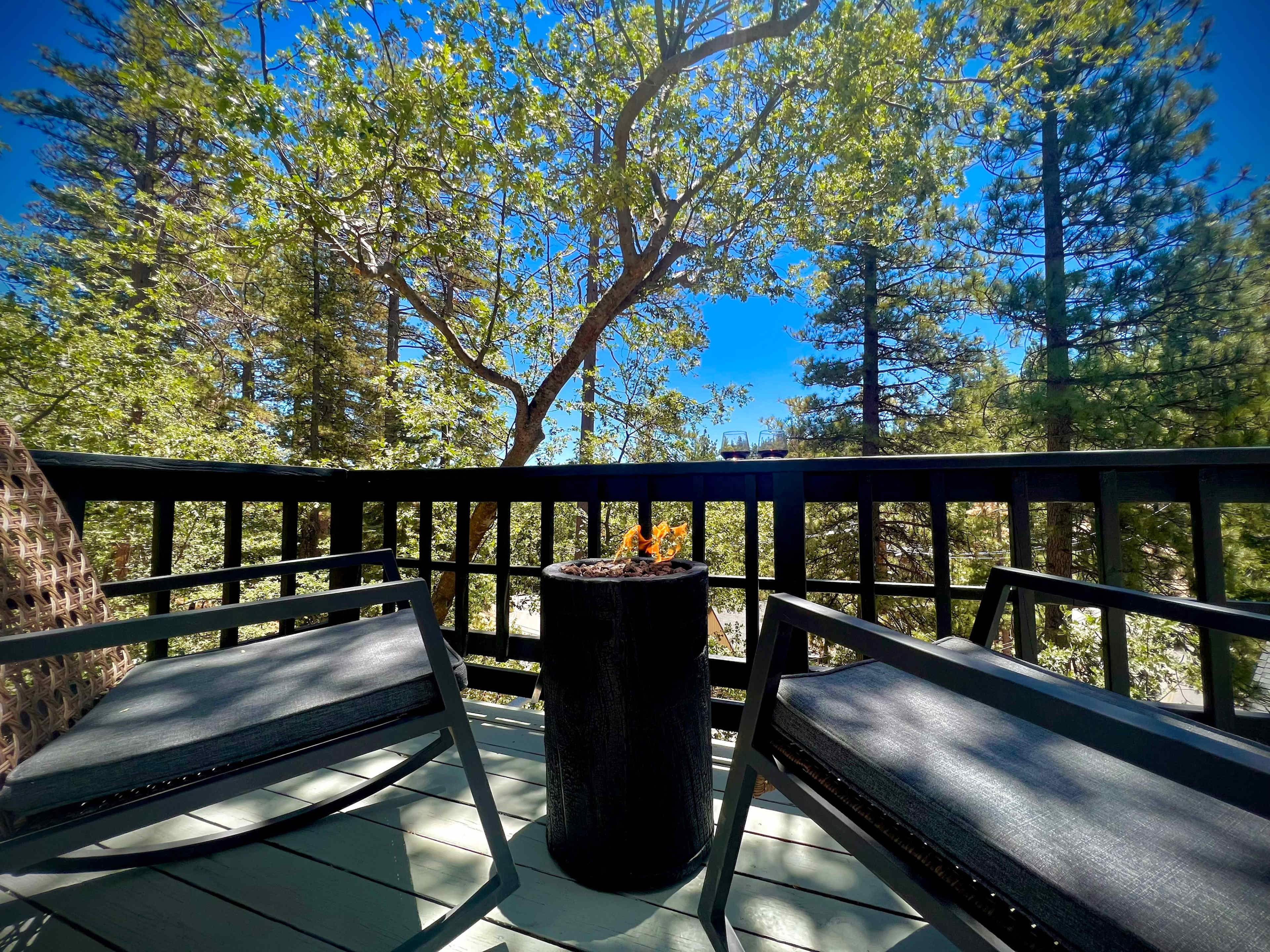 A deck with two black chairs and a small fire pit is surrounded by trees under a clear blue sky.