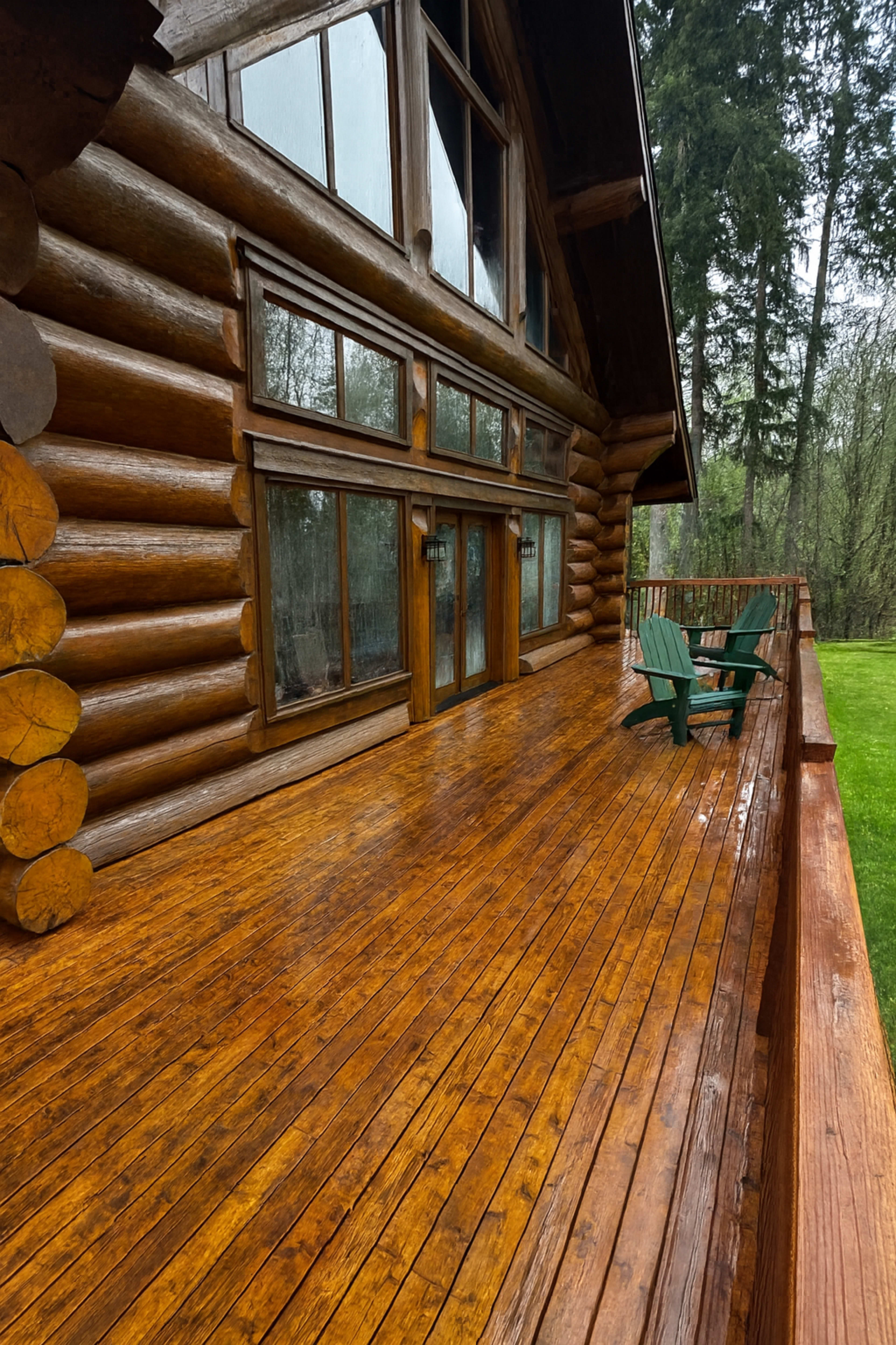 The image shows a wooden deck in front of a log cabin, featuring large windows and two green Adirondack chairs.