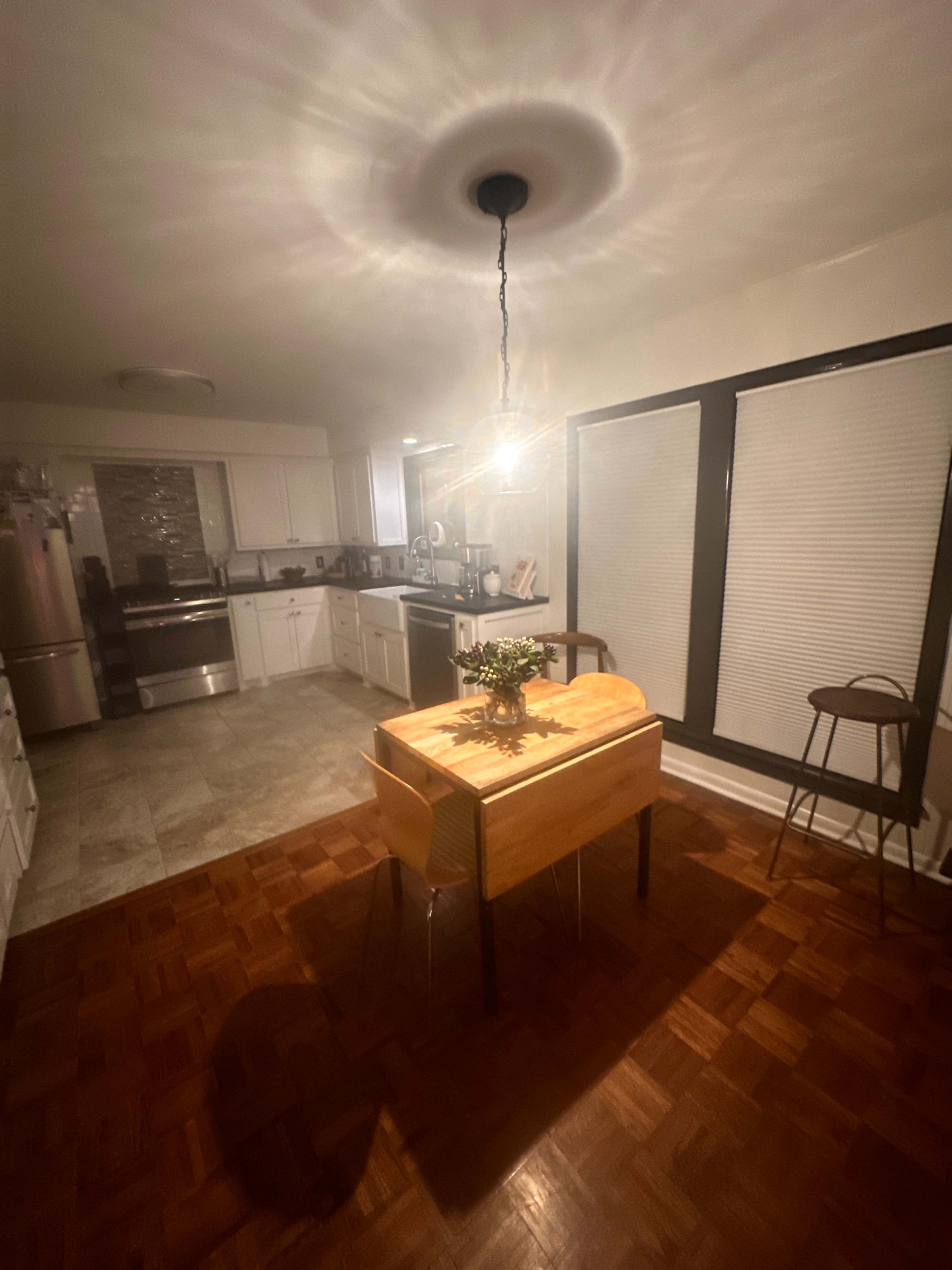 A small kitchen with a wooden table and a single hanging light illuminates the space, featuring a modern design with stainless steel appliances and large windows with blinds.