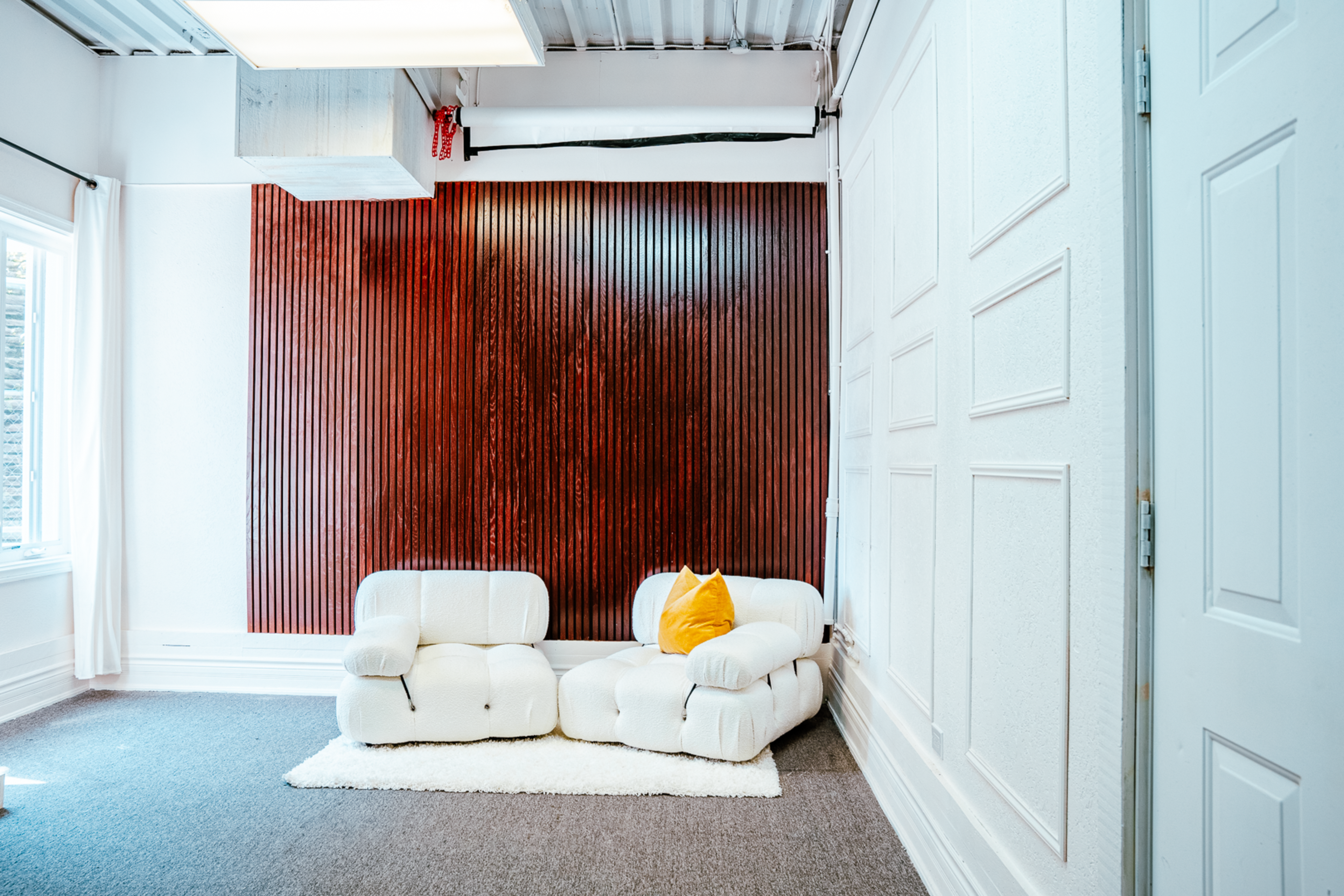 The image shows a minimalist room featuring a textured wooden wall and a white, modular sofa accompanied by a yellow pillow.