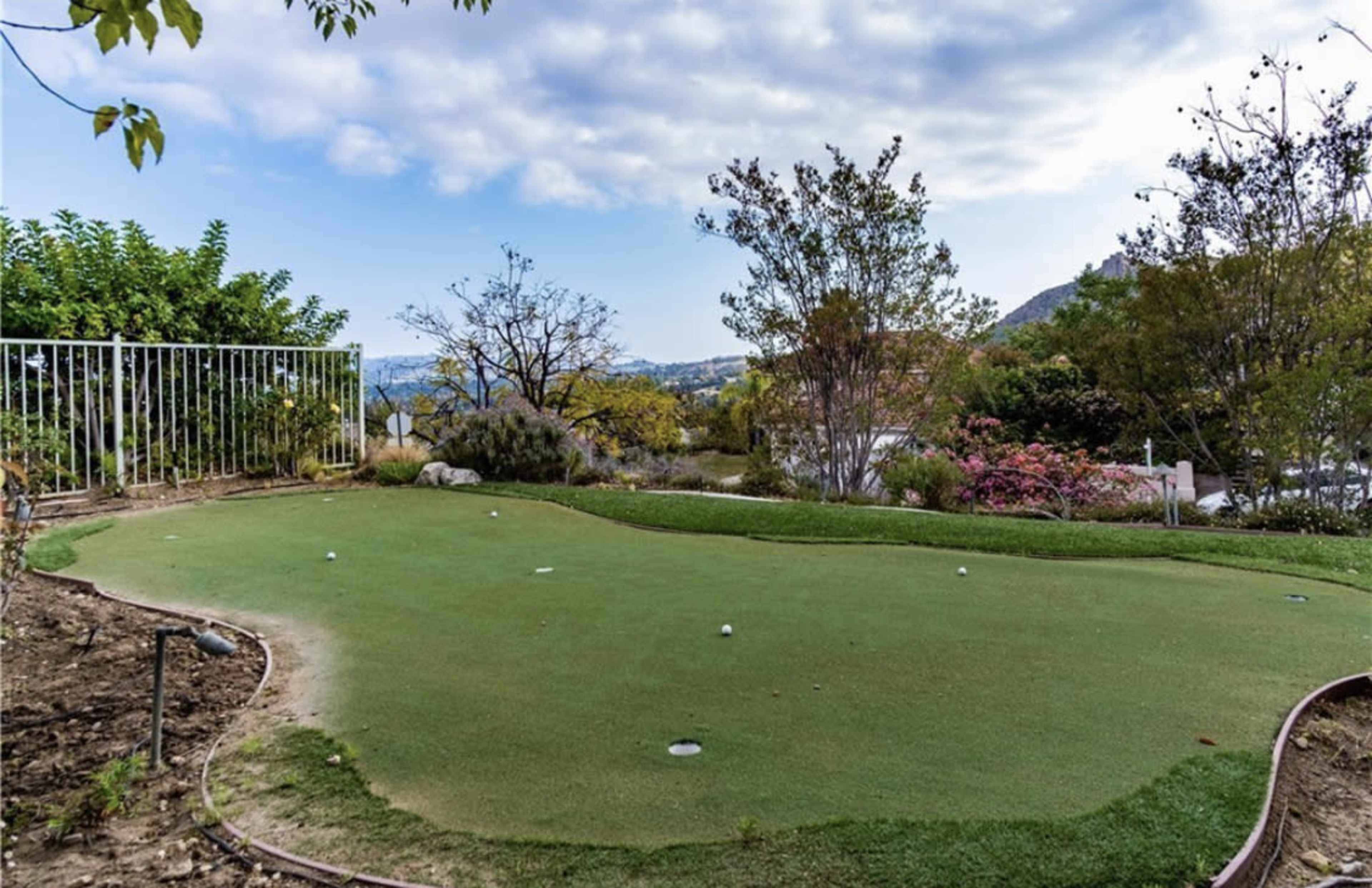 The image shows a small outdoor putting green surrounded by landscaped greenery and a view of distant hills.