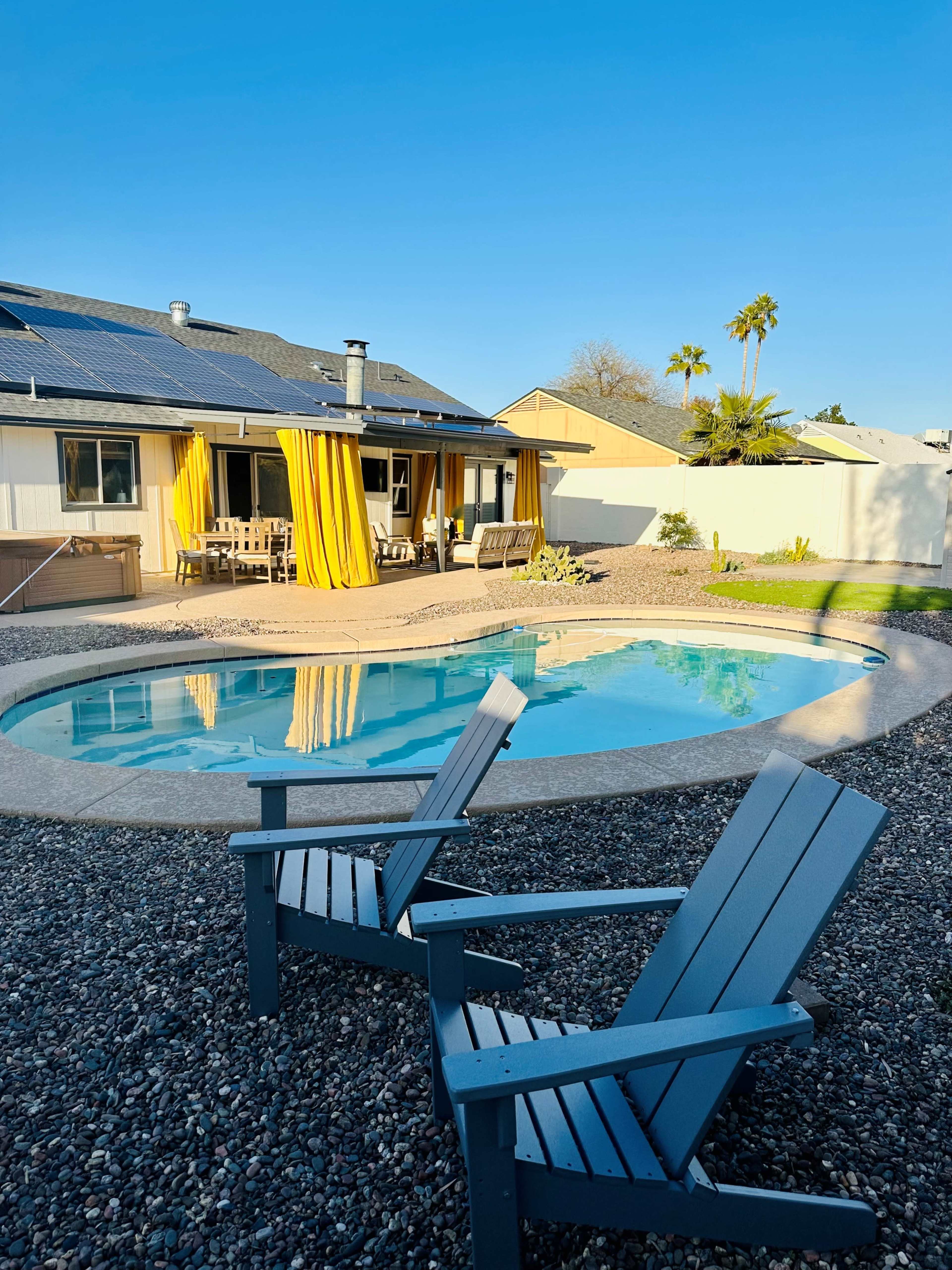A blue swimming pool is surrounded by gravel and two gray adirondack chairs, with a house featuring solar panels and yellow drapes in the background.