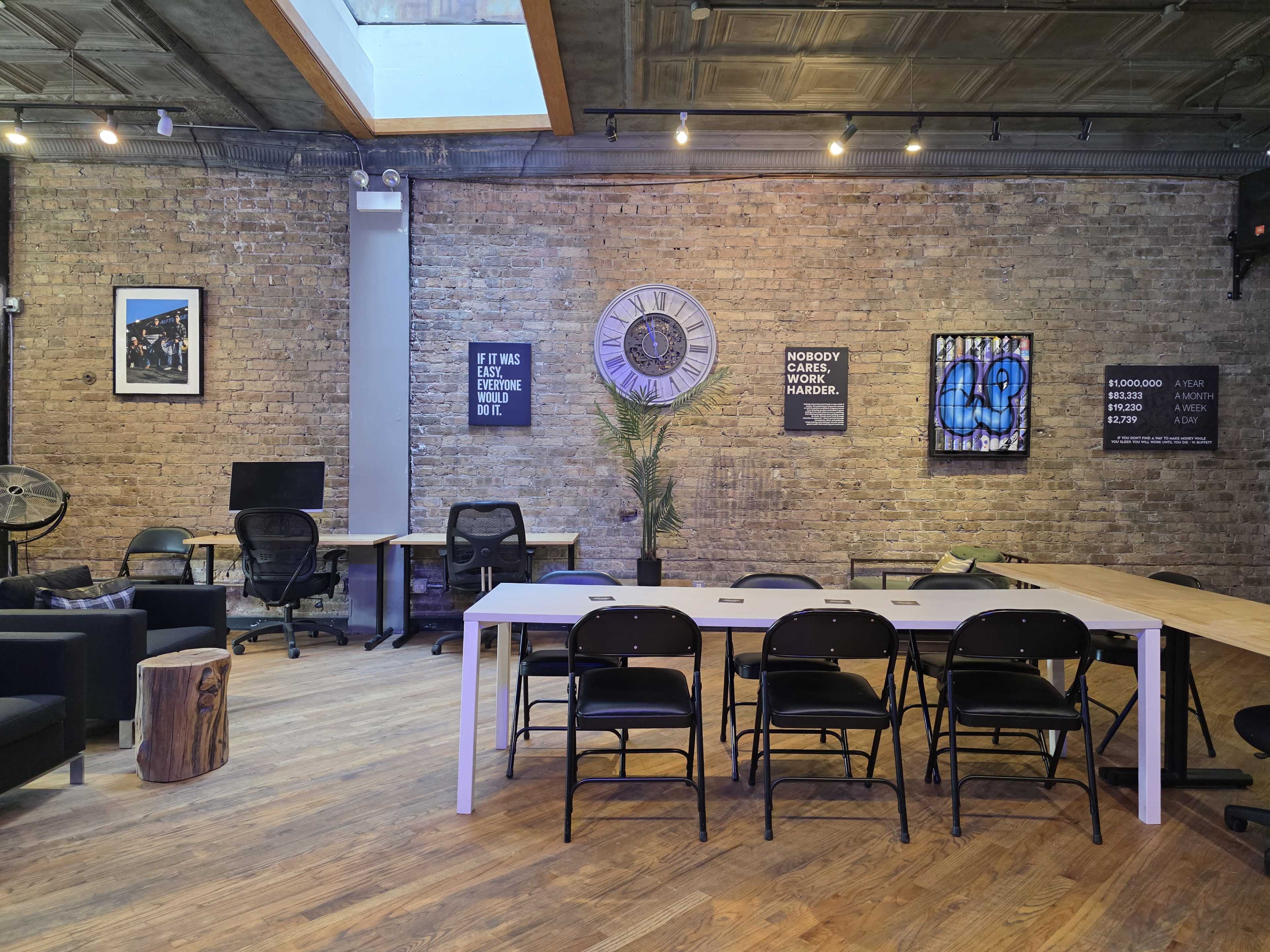 The image shows a modern office space with exposed brick walls, featuring a long table surrounded by black chairs, a large clock, and various artworks.